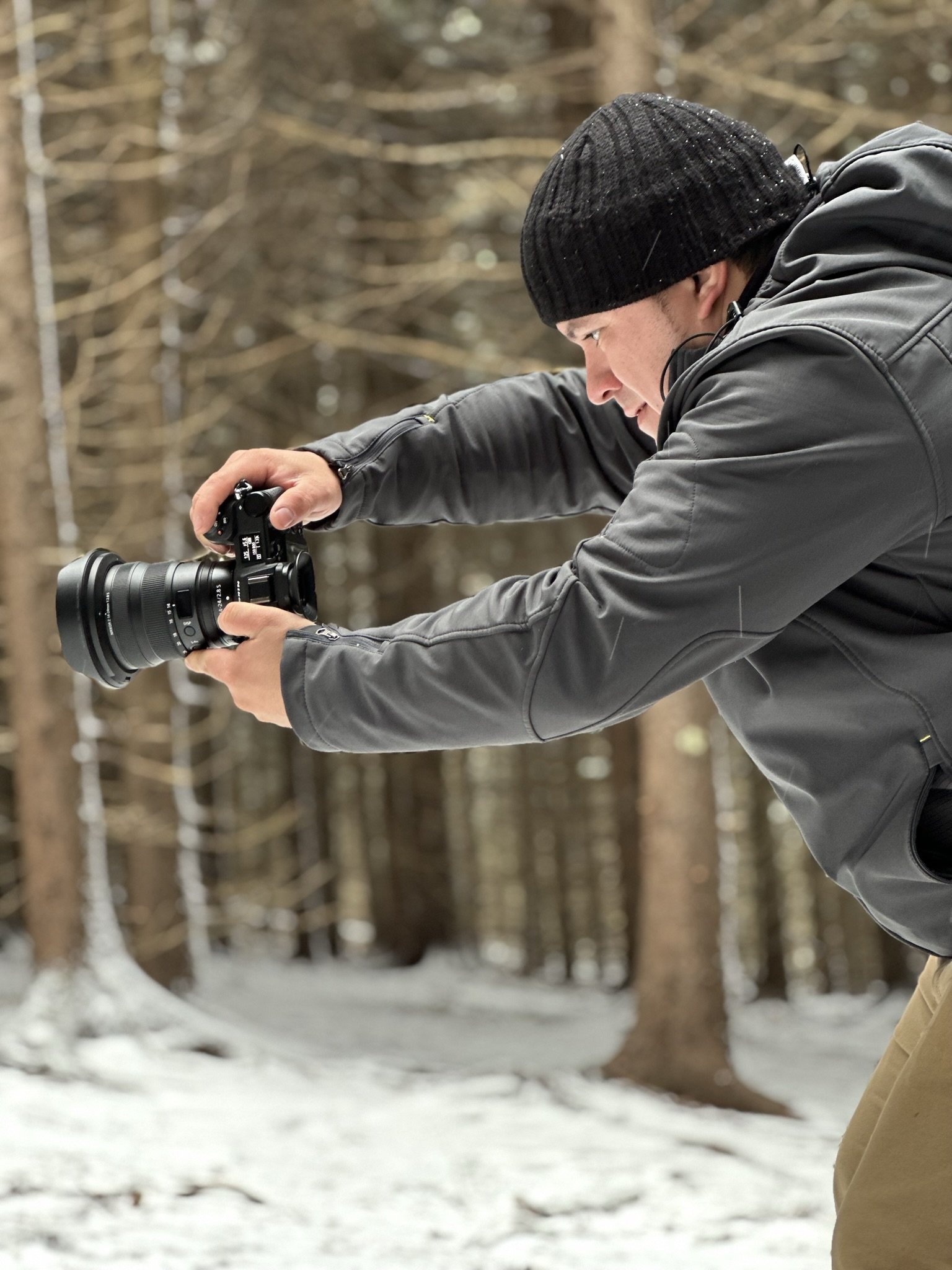 Melser Bonilla capturing a photo in a snowy forest, wearing a black beanie and gray jacket, focused on his camera.