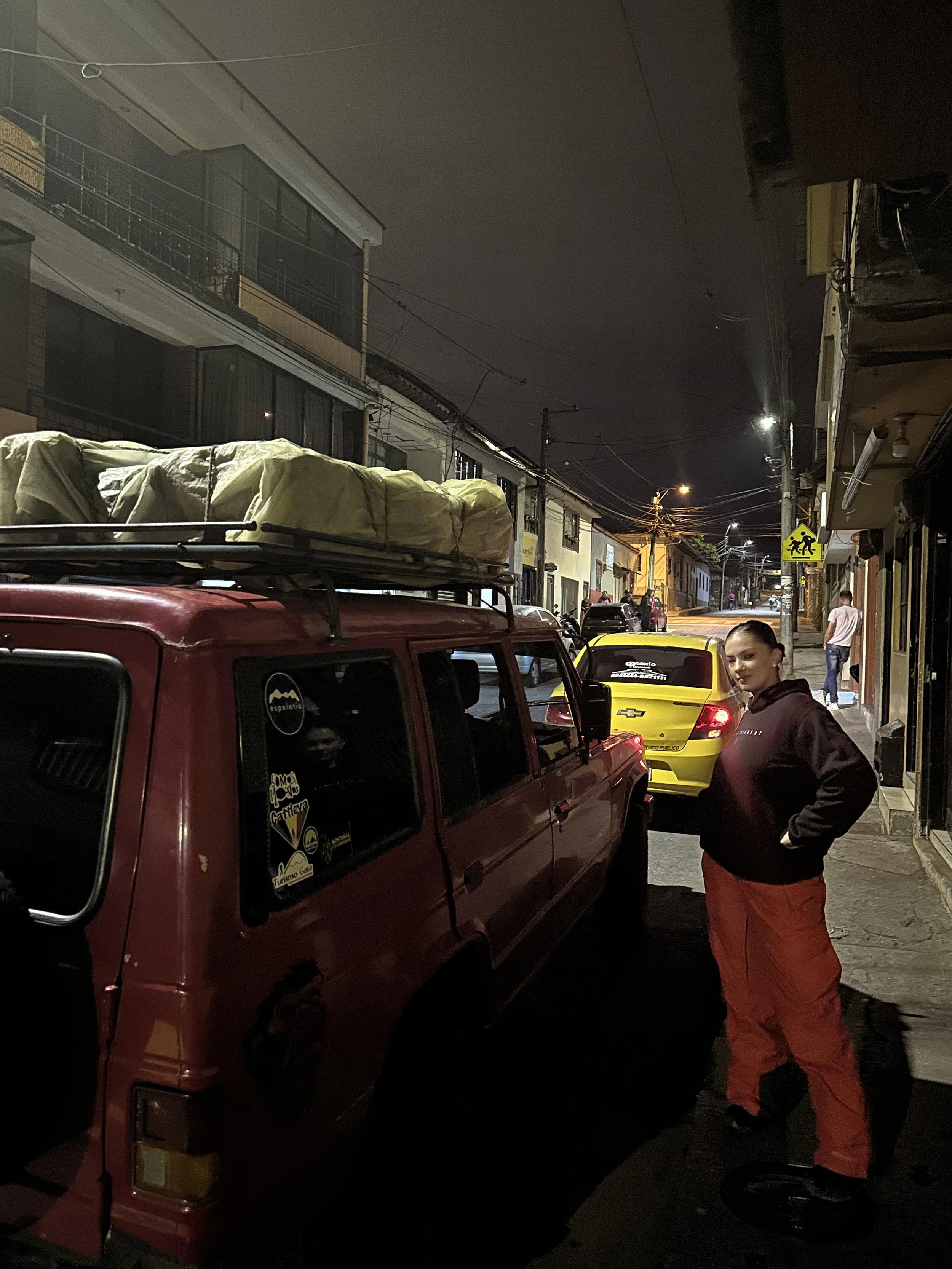 A woman standing next to a red pickup truck on a city street at night, with a yellow car behind her and buildings on both sides.