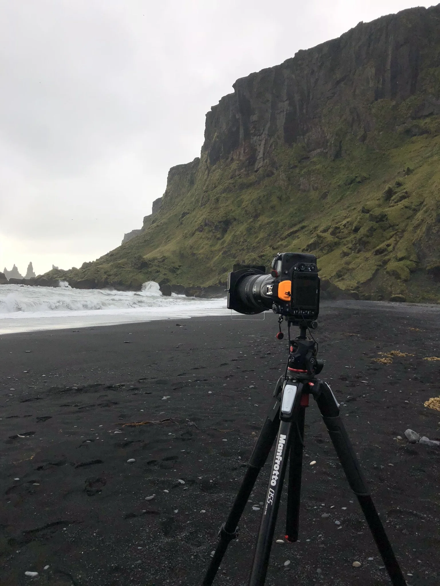 Camera on tripod facing the black sand beach with green mossy cliffs in the background and overcast sky.