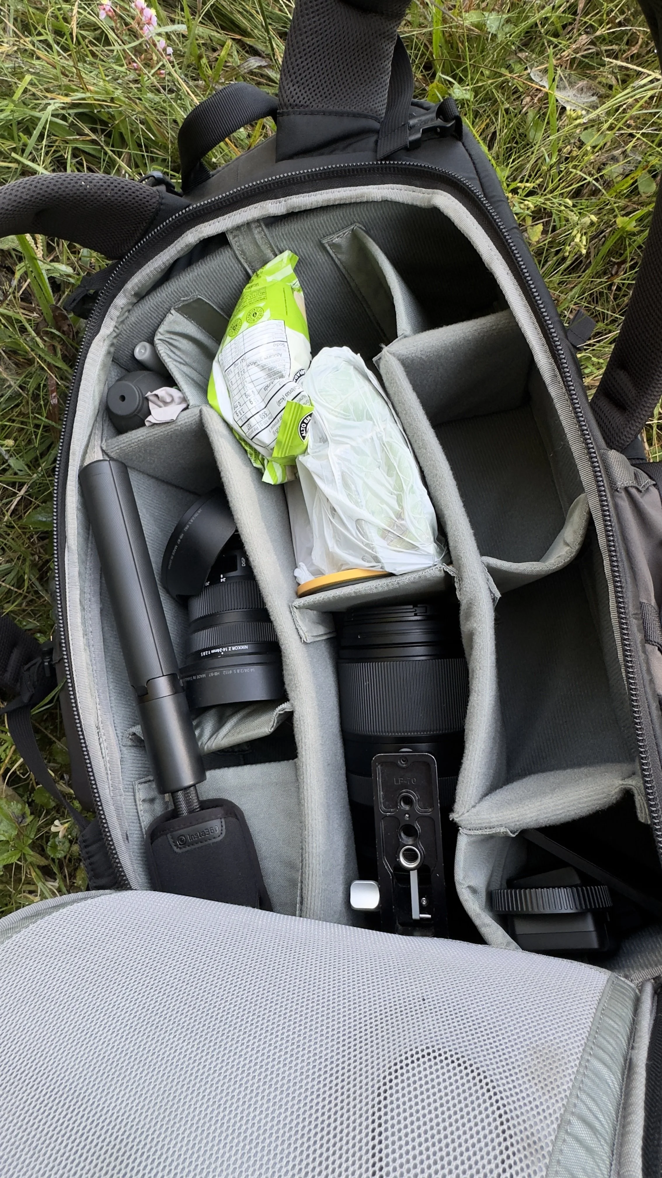 Camera bag with camera lens, a tripod, snacks, and other accessories inside, placed on grass.