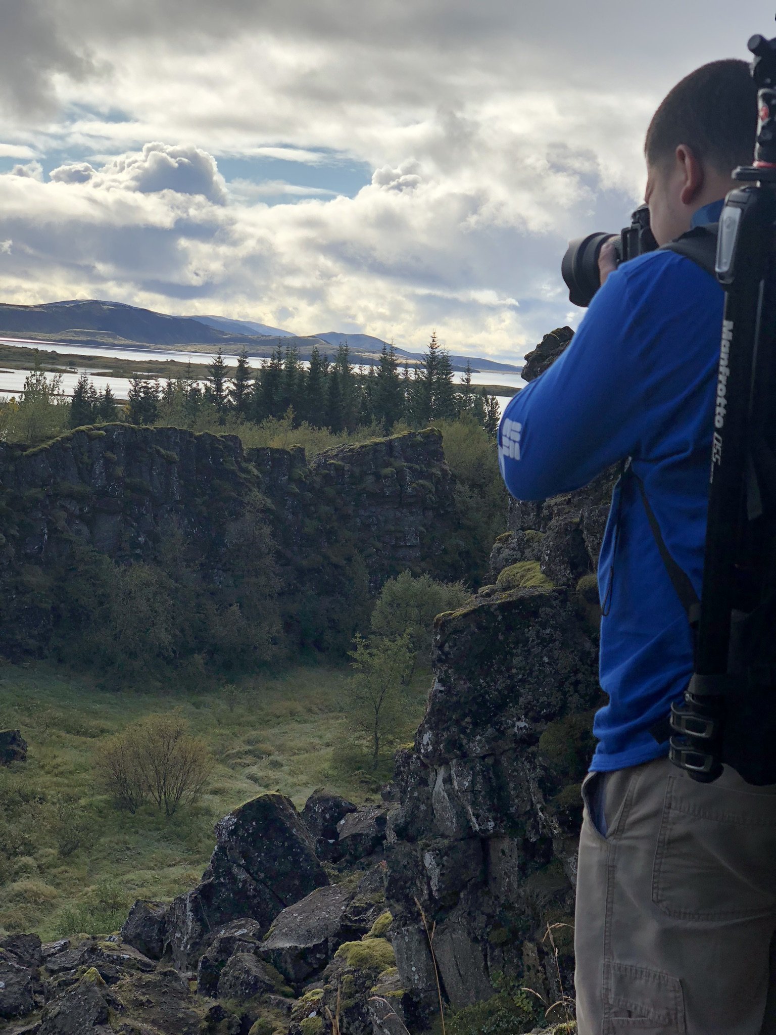 A man with a backpack taking a photograph of a scenic landscape with rocks, trees, a body of water, mountains, and a cloudy sky.