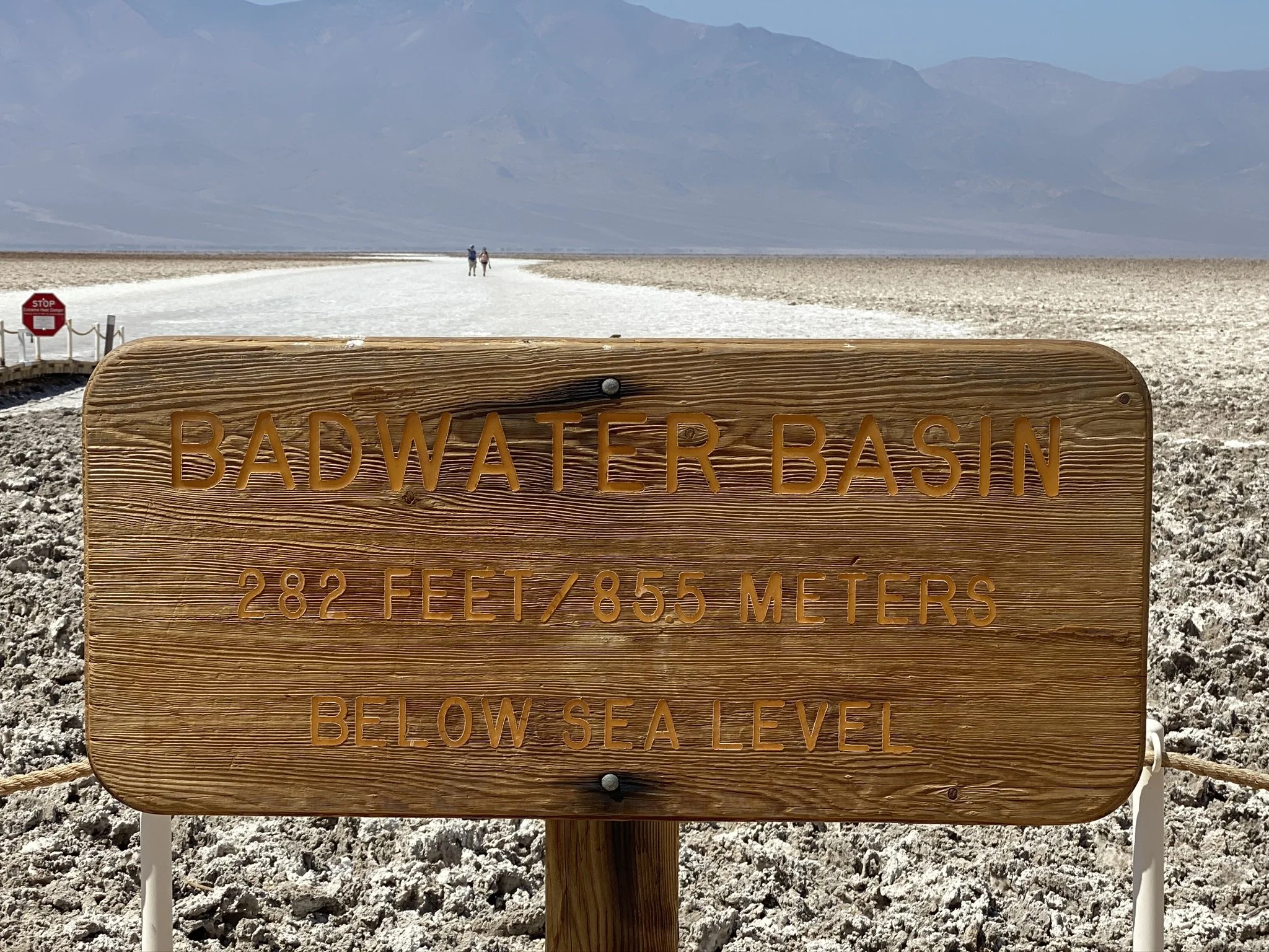 A wooden sign on a rope barrier in Badwater Basin, Death Valley, California, indicating an elevation of 282 feet below sea level. The background shows a salt flat landscape with two people walking in the distance, mountains and a cloudy sky.