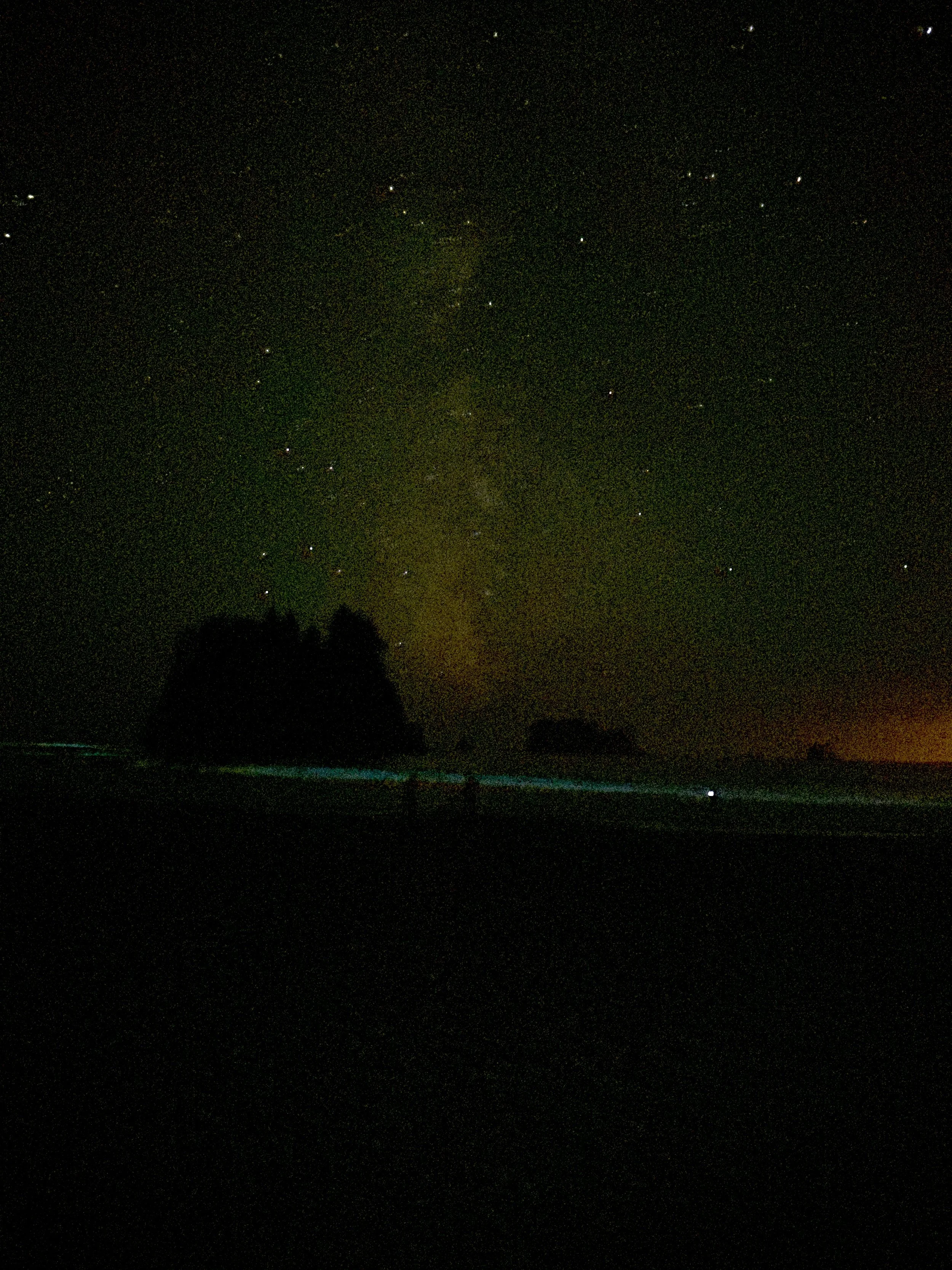 Nighttime image showing stars in the sky, silhouettes of trees or rocks on the horizon, and a faint glow near the horizon, possibly from city lights or sunset.
