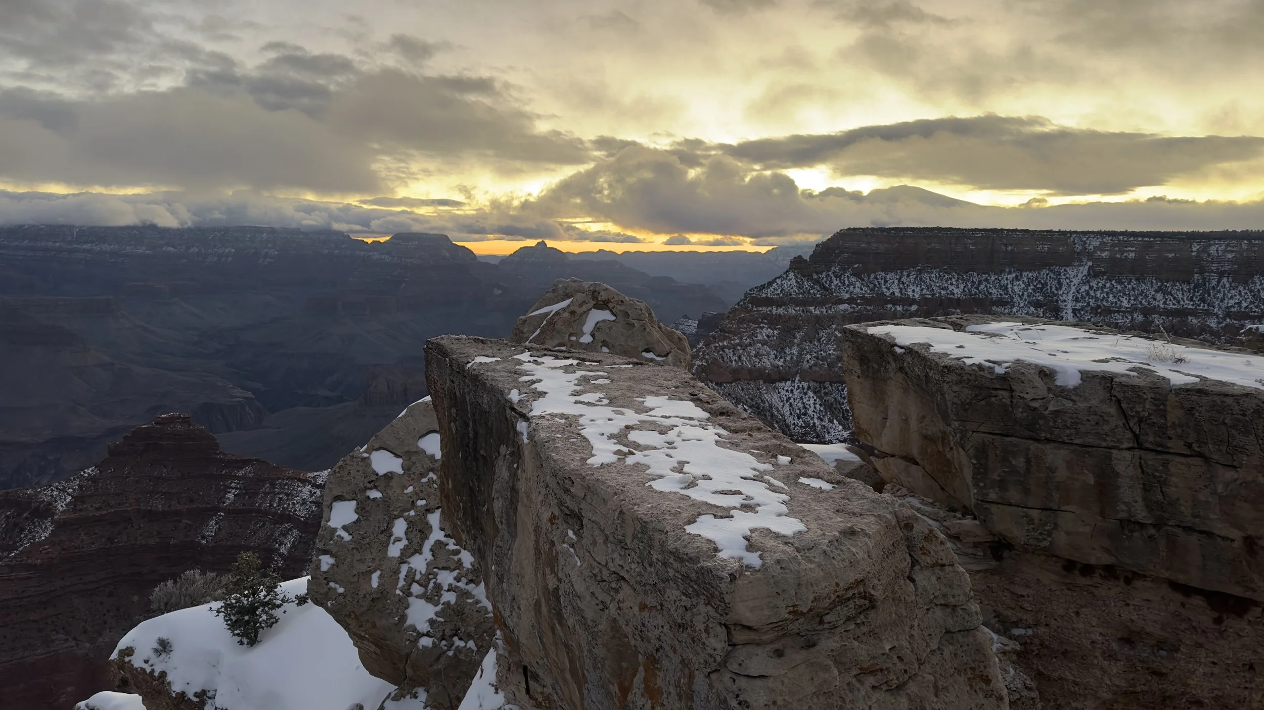 Snow-dusted rock formations and the Grand Canyon in the distance during sunset with cloudy skies.