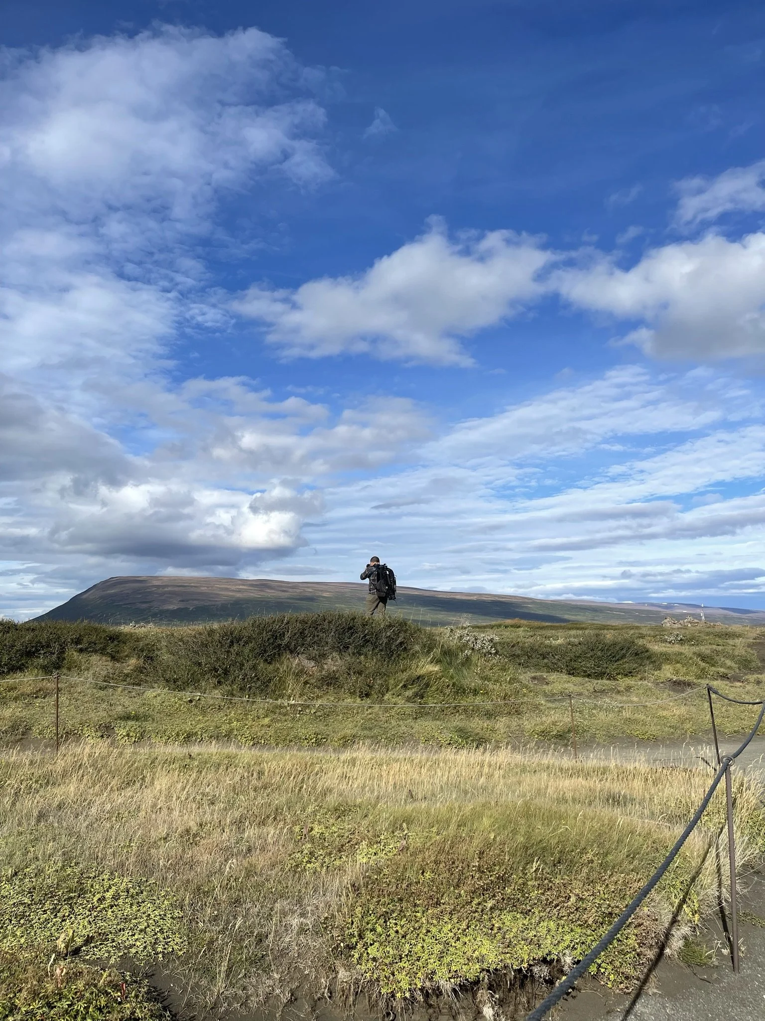 A person with a backpack standing on grassy terrain under a partly cloudy sky, with a mountain in the background.