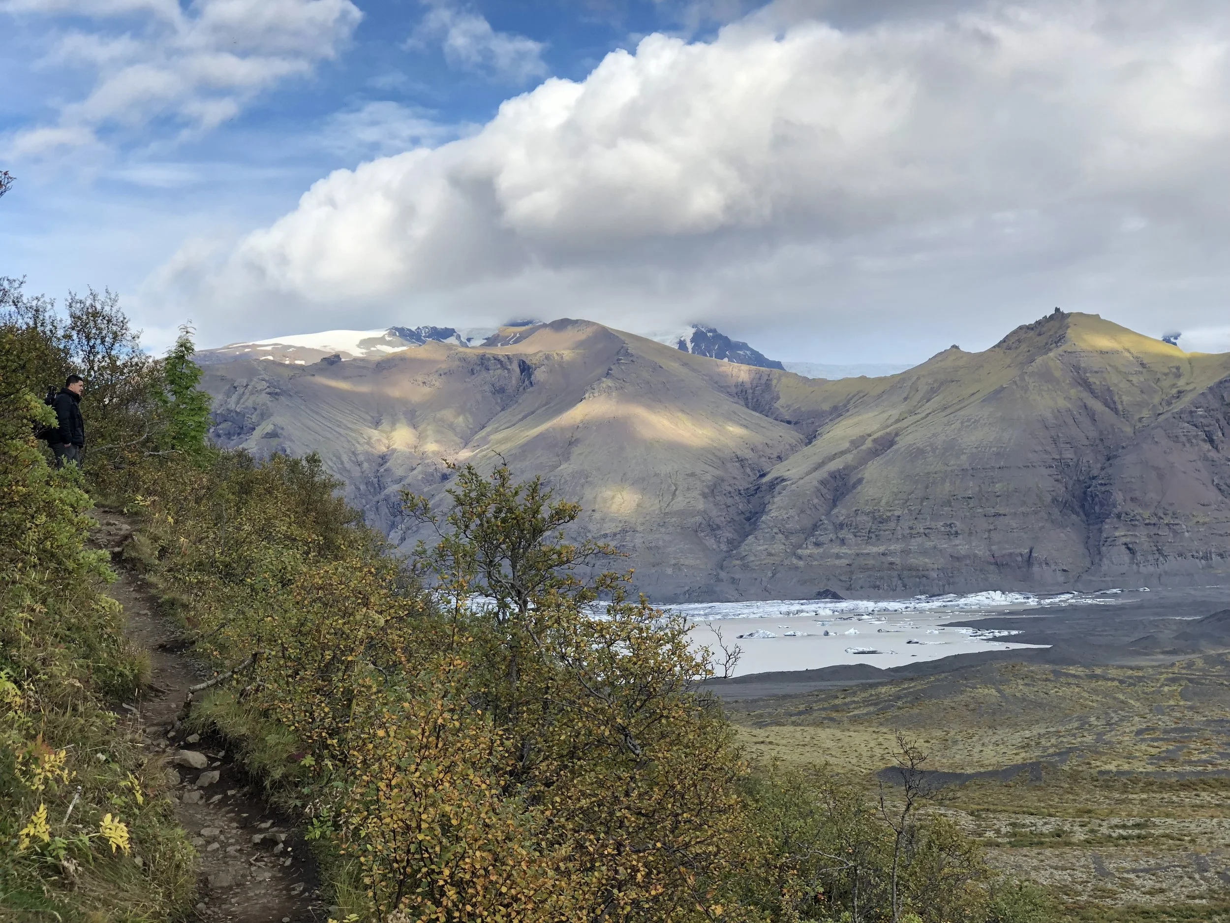 A hiker walking on a trail in a mountainous landscape with snow-capped peaks, green vegetation, and a partly cloudy sky.