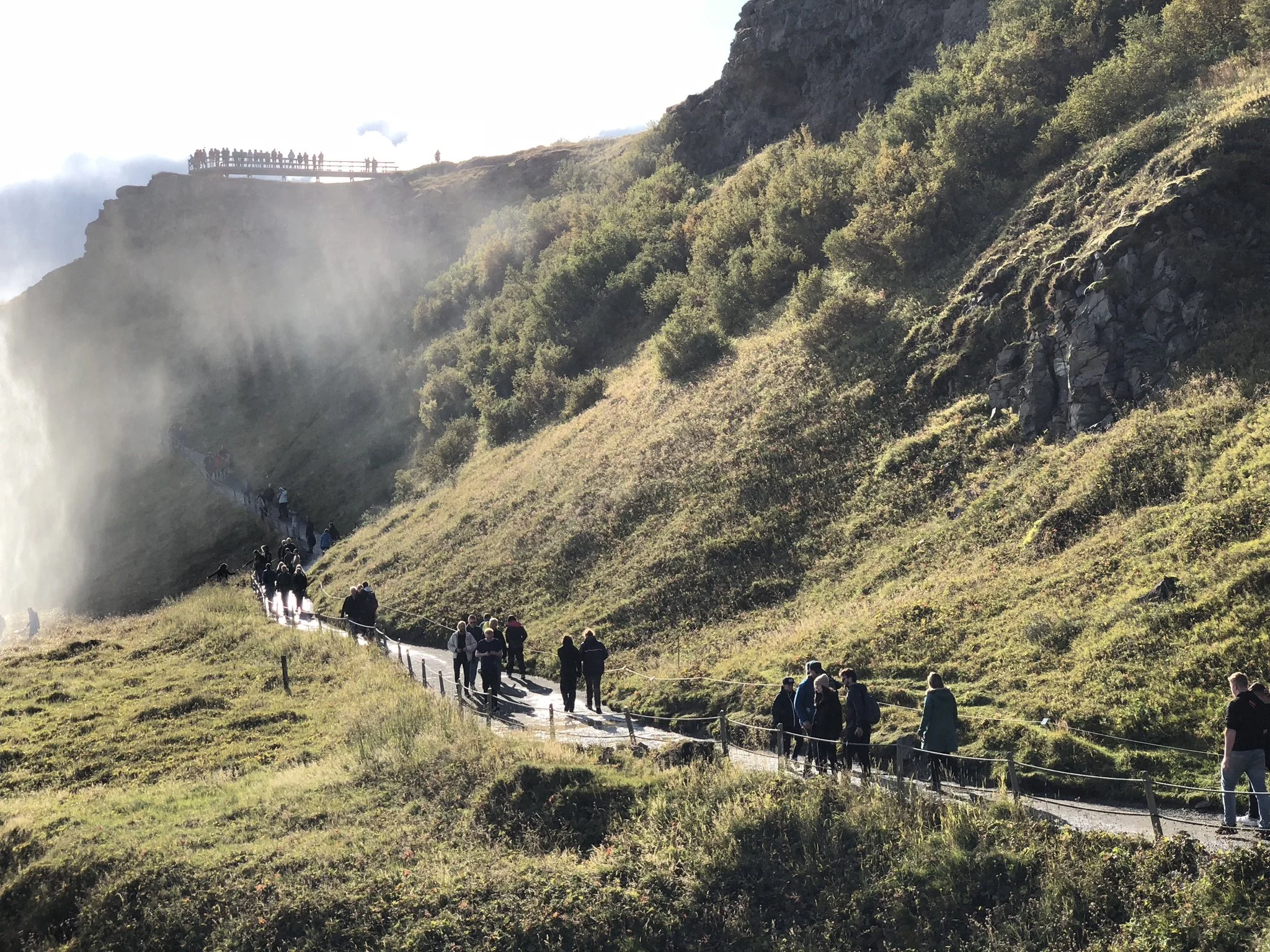 People walking along a trail through a lush, green mountainous landscape with mist and sunlight, leading to a viewpoint on a cliff.