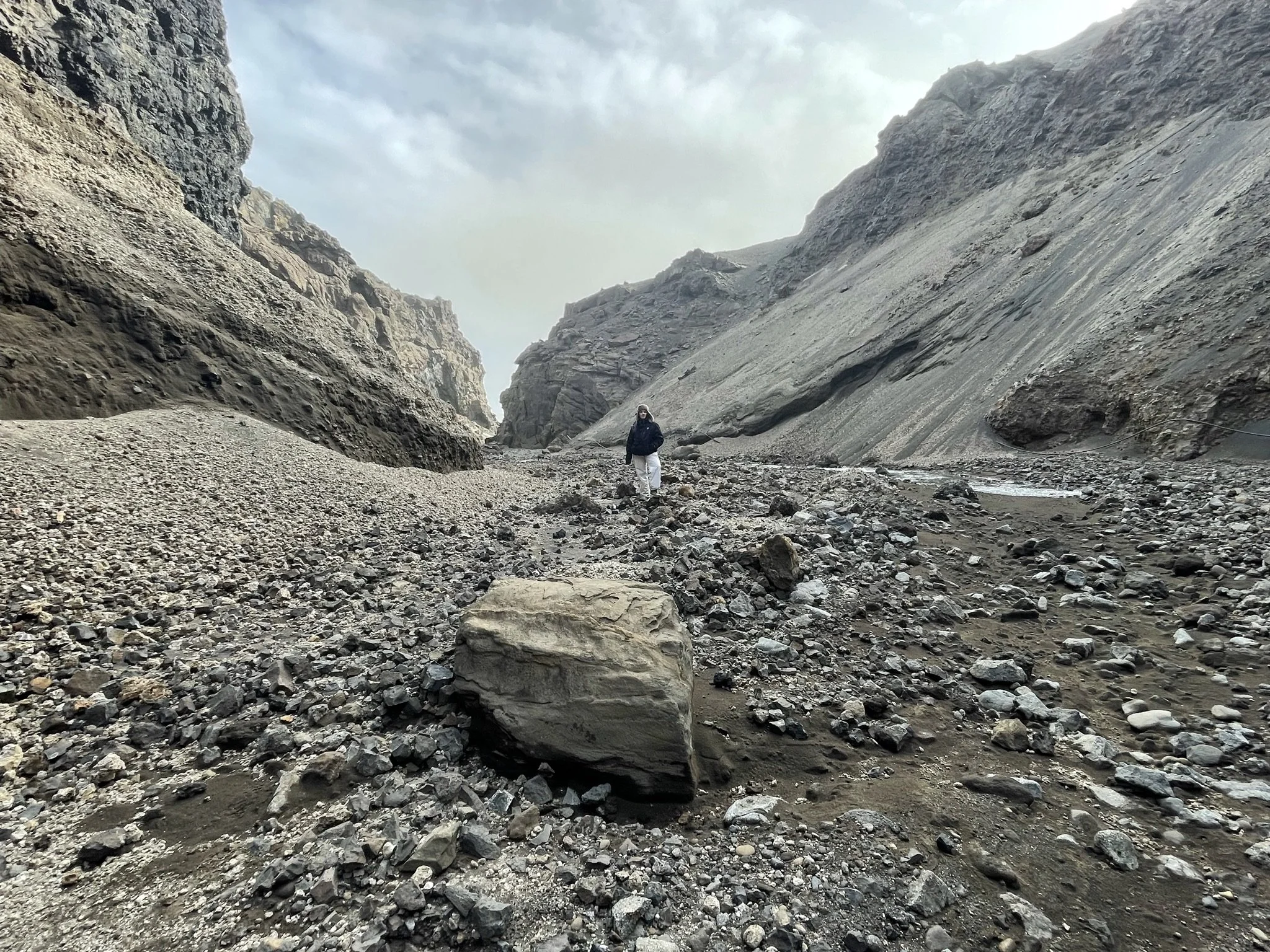 A person stands in a rocky canyon with steep, layered cliffs on both sides and a cloudy sky above.