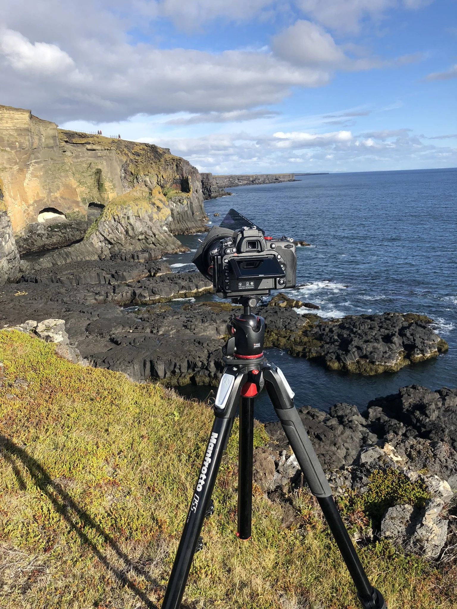 A camera mounted on a tripod set up on a grassy cliff overlooking the ocean with rocky shoreline and cliffs in the background.
