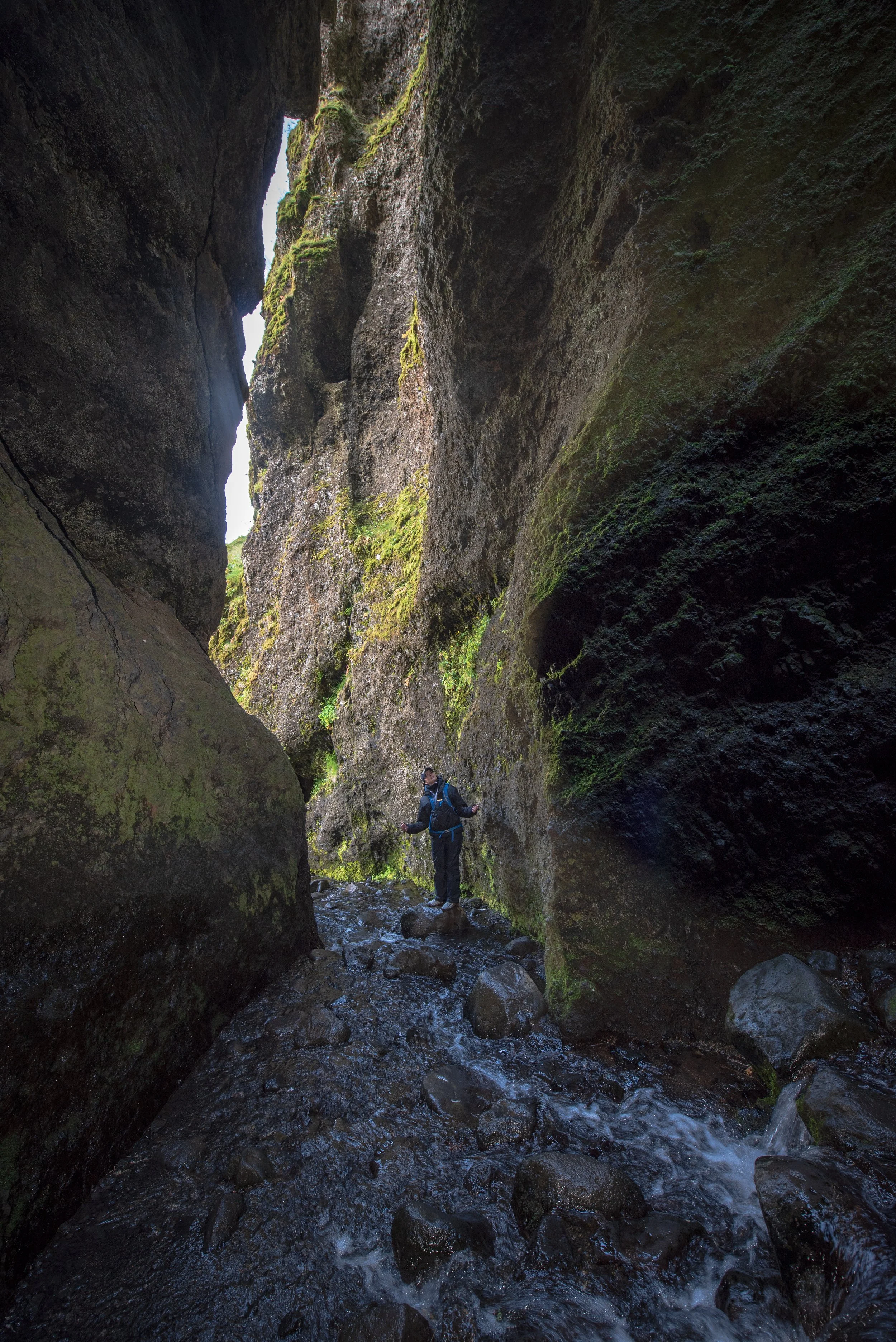 A person hiking through a narrow, moss-covered canyon with water flowing on the rocky ground.