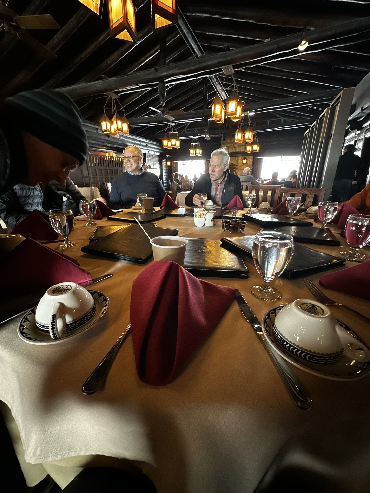 A dining table set with red napkins, teacups, glasses of water, and menus in a cozy restaurant with wooden interior and hanging lantern lights, with people seated and chatting in the background.