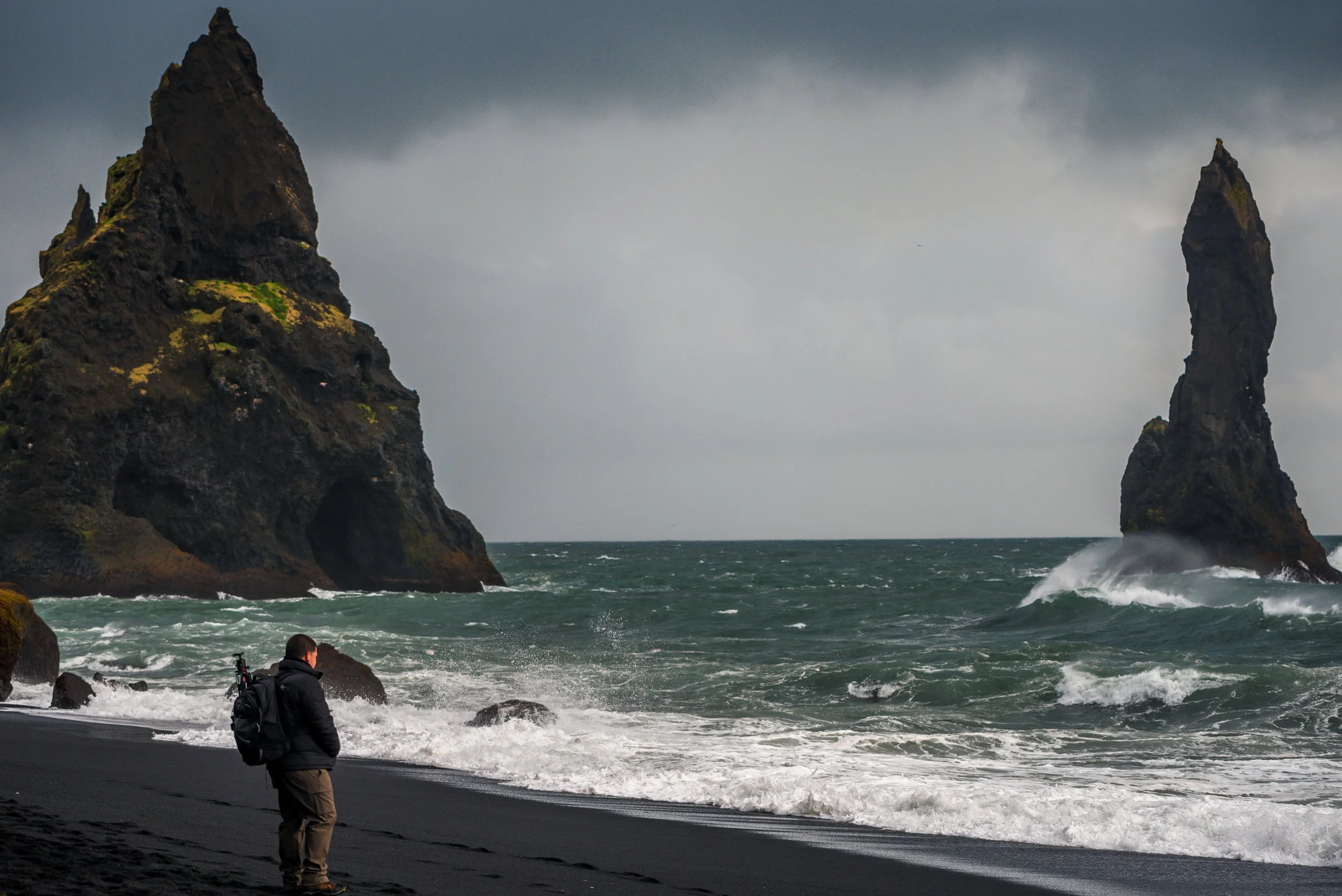 A person standing on a black sand beach with two large sea stacks in the ocean behind them, under a cloudy sky.
