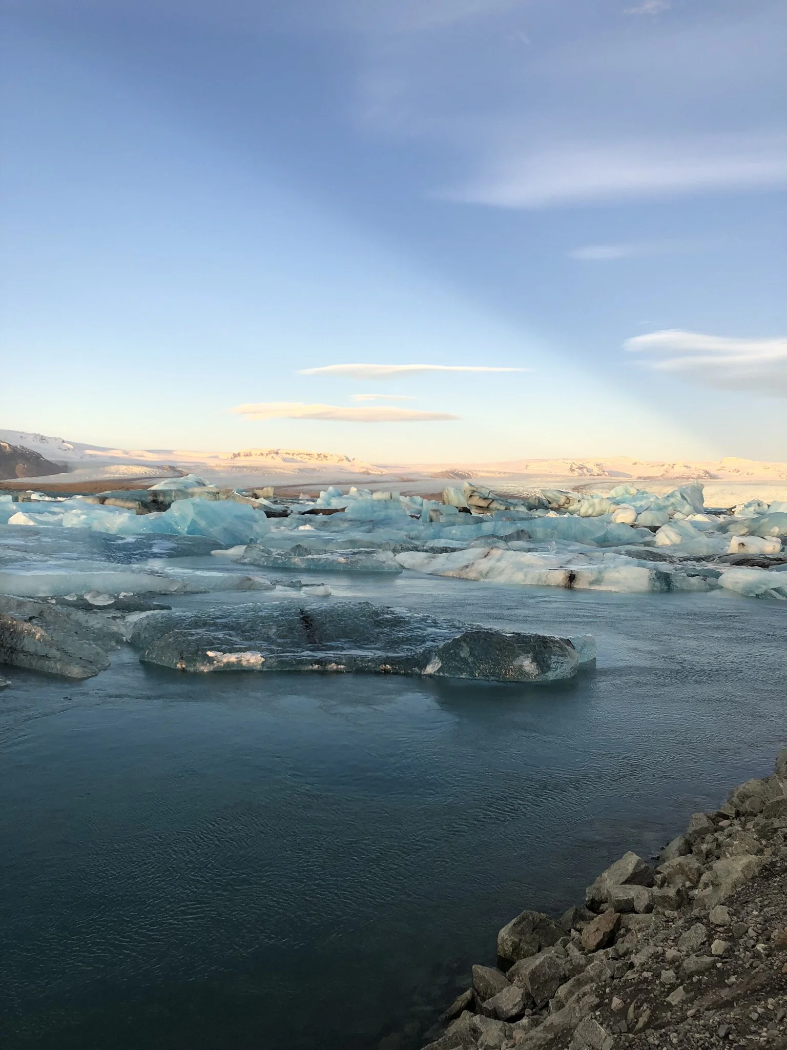 Icebergs floating in a cold, clear body of water with a rocky shoreline, mountains, and an expansive sky with light clouds in the background.