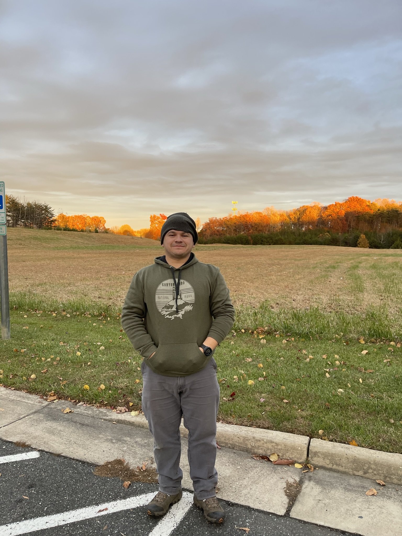 A man standing outdoors in a parking lot during autumn, wearing a black beanie, an olive green hoodie, gray pants, hiking shoes, with trees showing fall foliage in the background and an overcast sky.