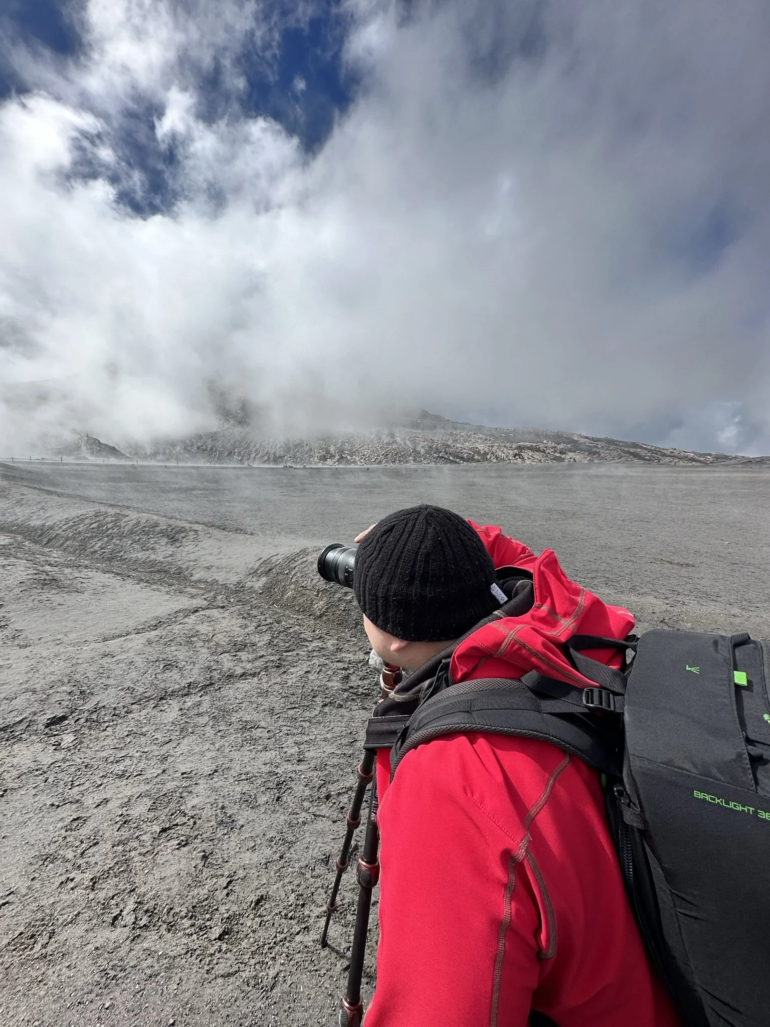 A person wearing a black beanie and red jacket taking a photo with a camera on a tripod in a volcanic landscape with gray ash and steam, cloudy sky overhead.