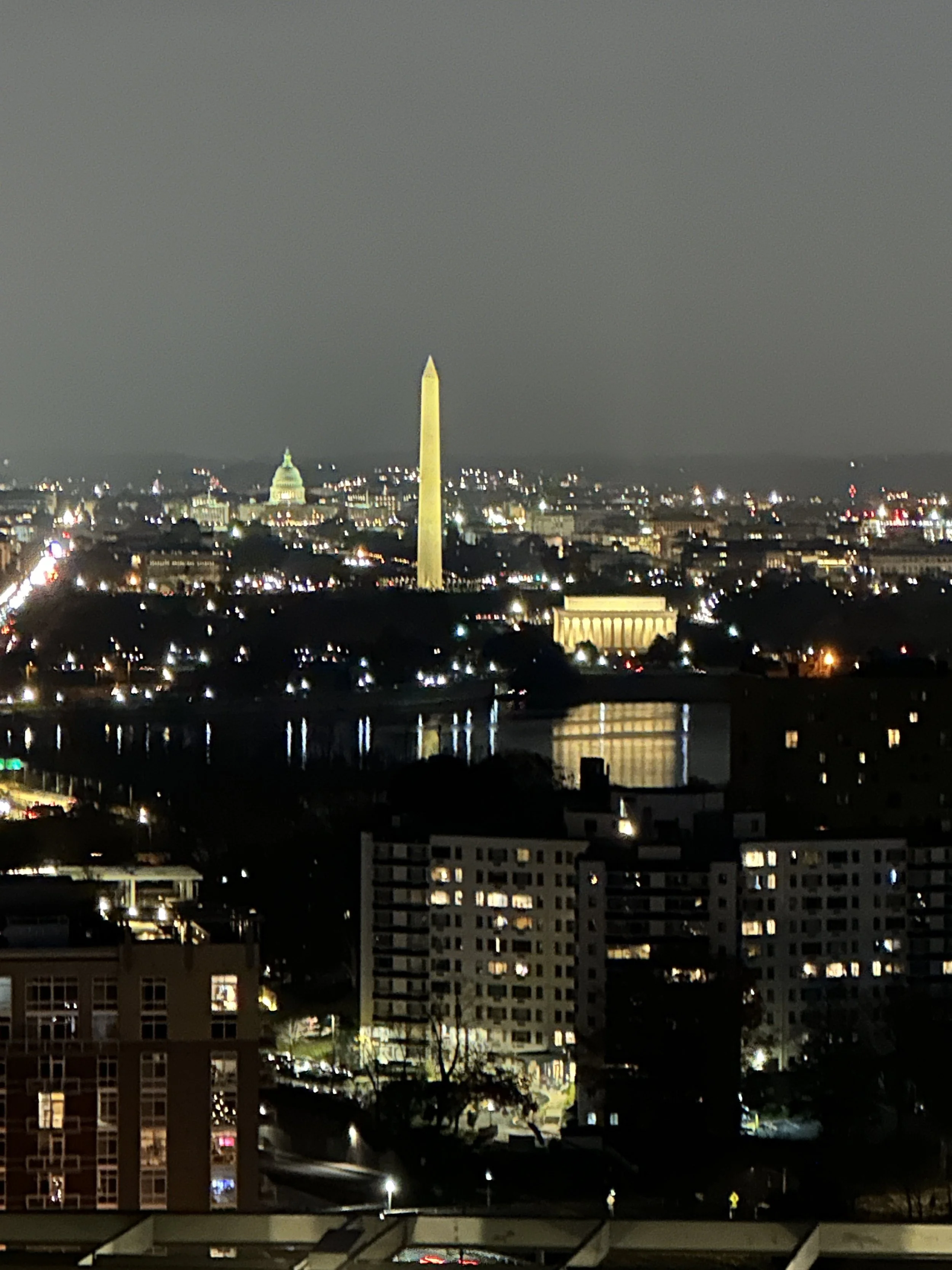 Nighttime cityscape of Washington, D.C., featuring the illuminated Washington Monument, the Lincoln Memorial, and the U.S. Capitol building in the background, with reflections on the water and lit buildings in the foreground.