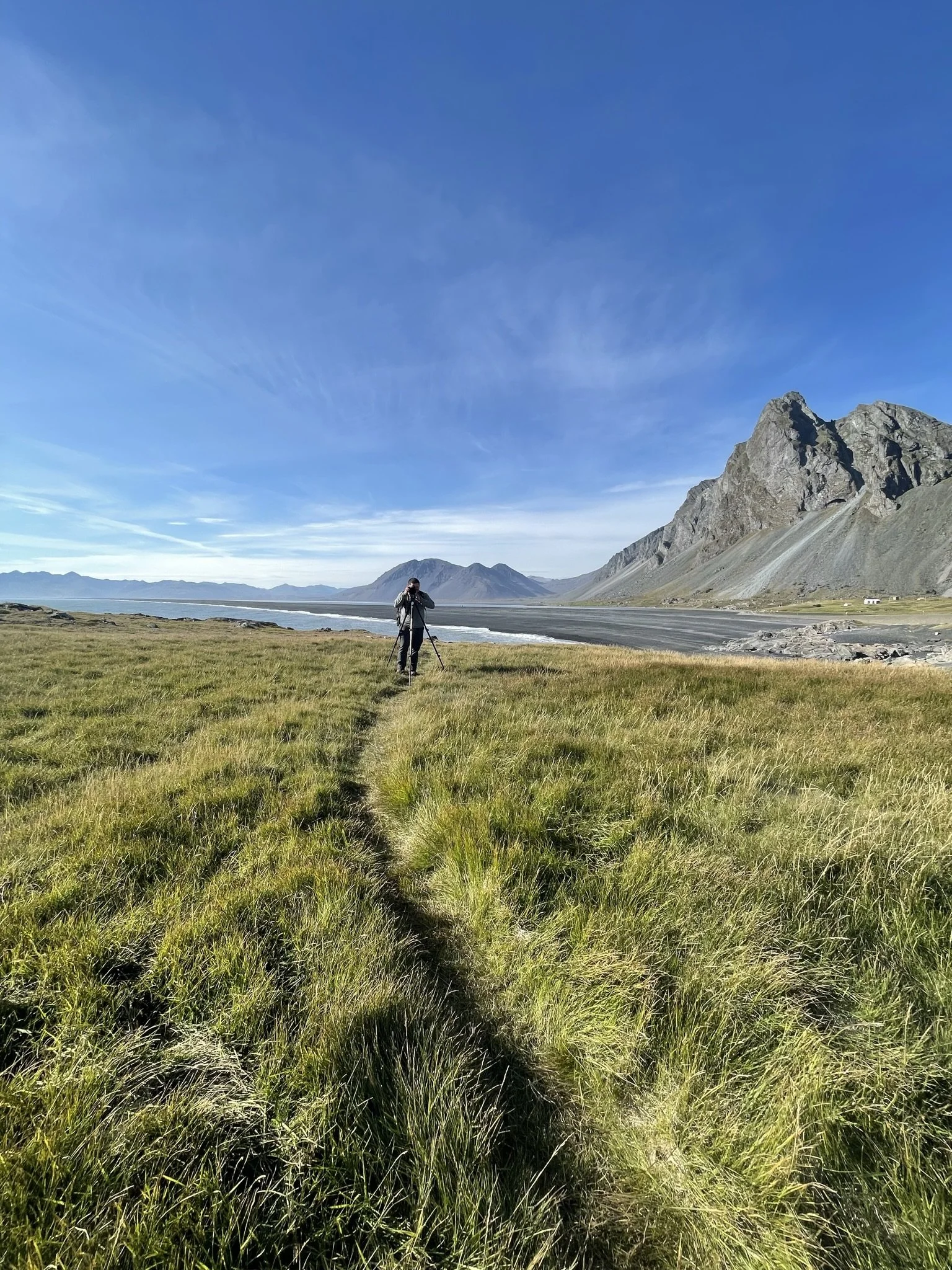 A person with a camera on a tripod walking along a grassy trail towards a body of water with mountains in the background on a sunny day.