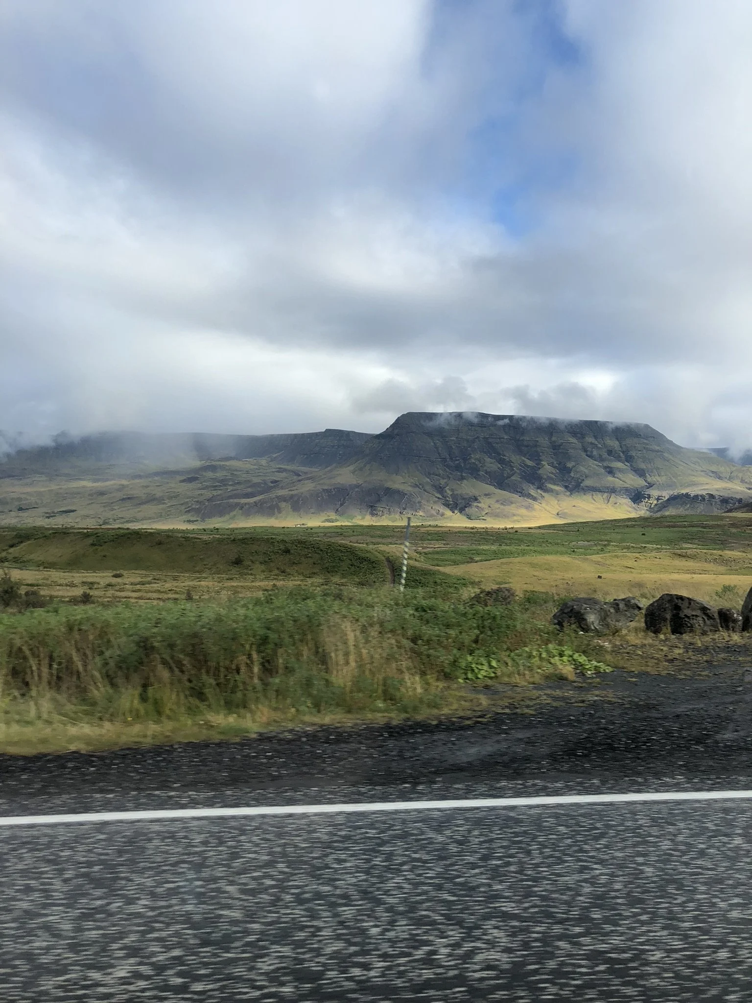 Scenic view of a mountain range in Iceland with cloudy sky, green fields, and rocky terrain, taken from a moving vehicle on a highway.
