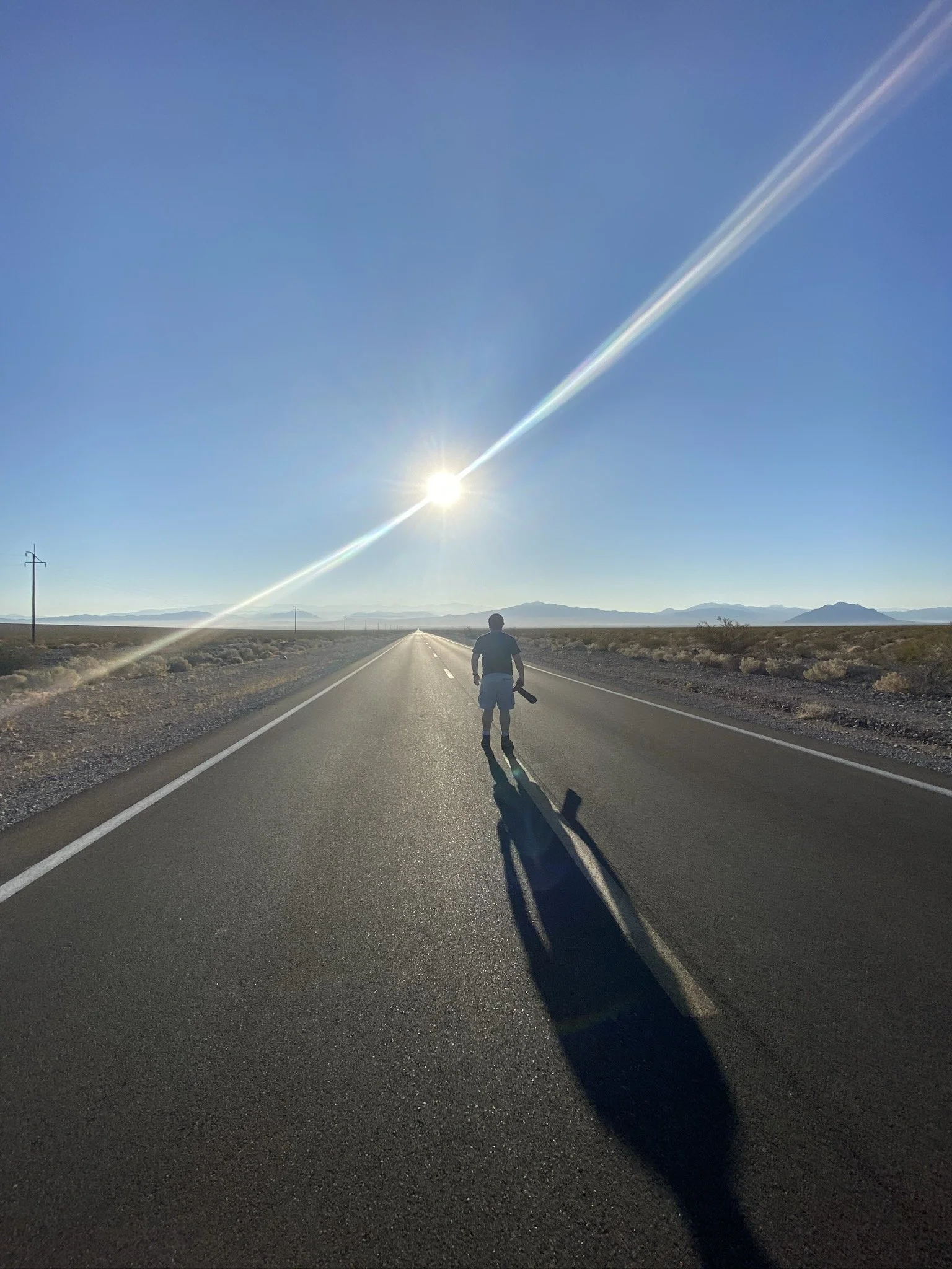 A person walking on a deserted highway in a desert area with mountains in the distance under a bright, clear sky and the sun shining overhead.