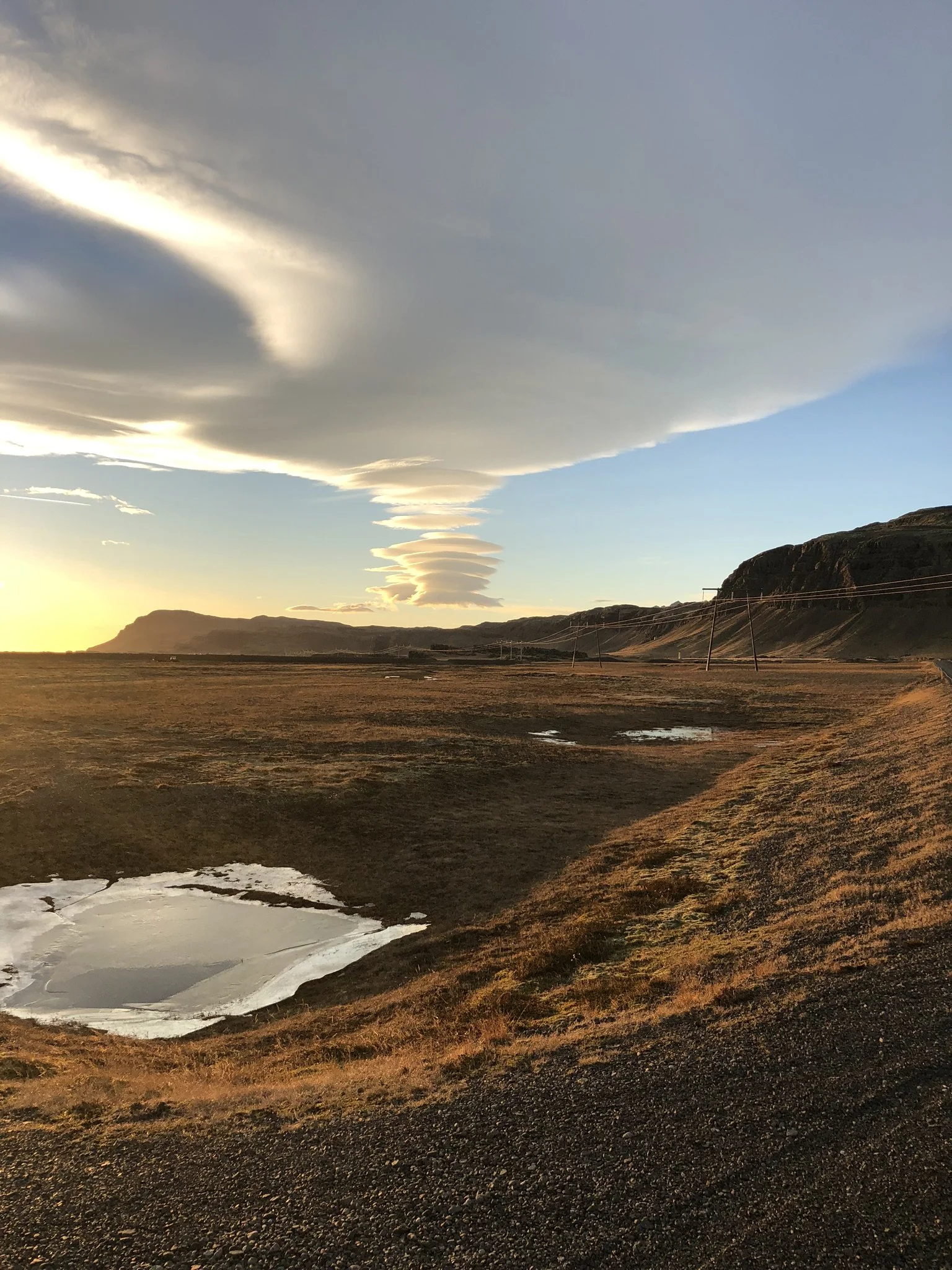 A landscape scene at sunset with a large, layered cloud formation in the sky, hills in the distance, and a small pond in the foreground with patches of grass and water.