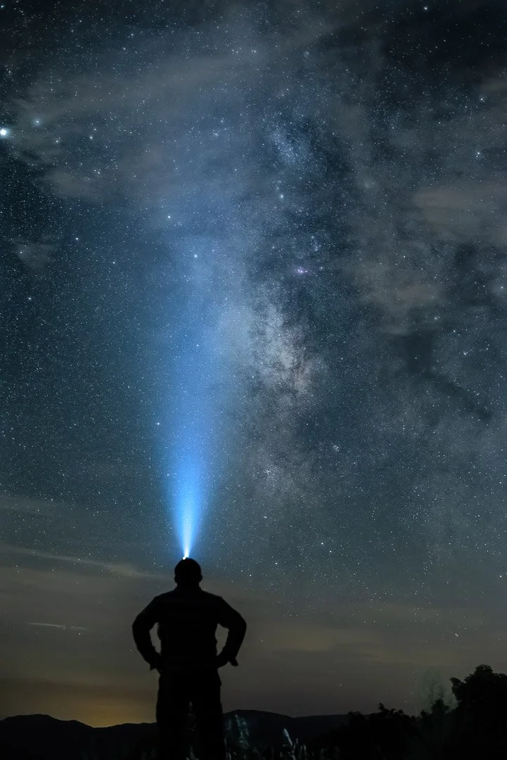 A person standing outdoors at night looking up at a starry sky with the Milky Way galaxy visible; the person is holding a flashlight directed till the sky.
