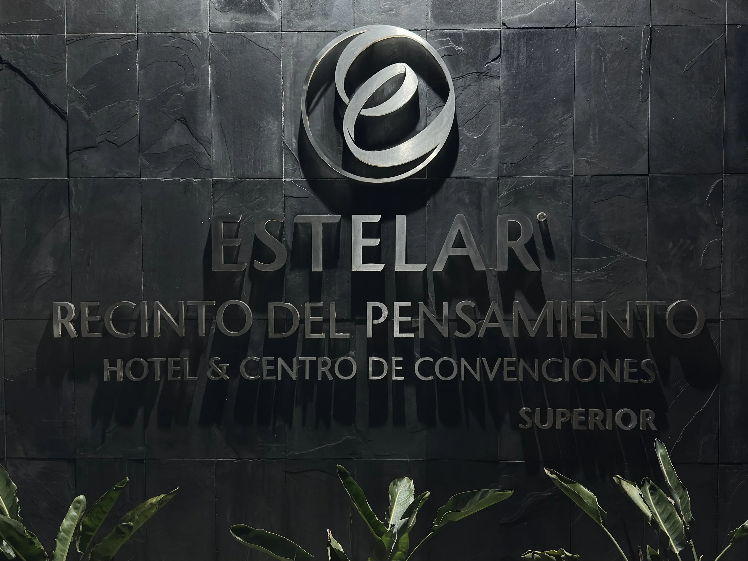 Sign for Estelari, a hotel and convention center, on a dark wall with metallic letters and a circular logo above. Green plants are at the bottom.
