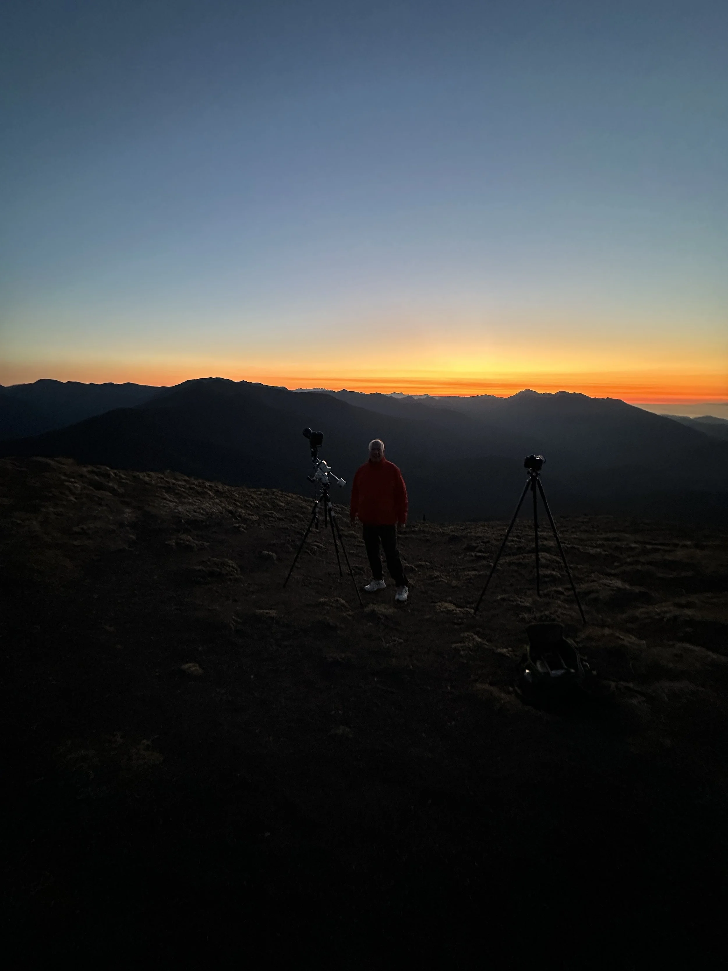 Person standing next to two tripods with cameras on a mountain at sunset, silhouette against colorful sky.
