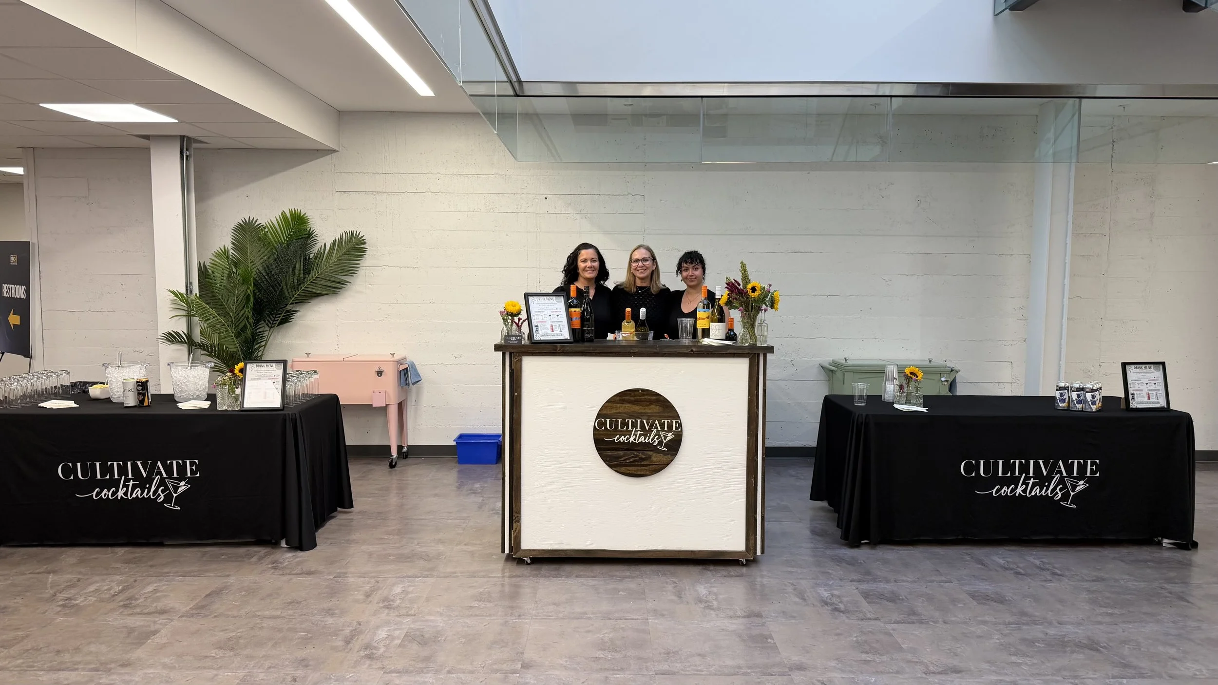 Three women behind a cocktail bar with drinks, flowers, and a menu, inside a modern venue with white walls and glass ceiling.