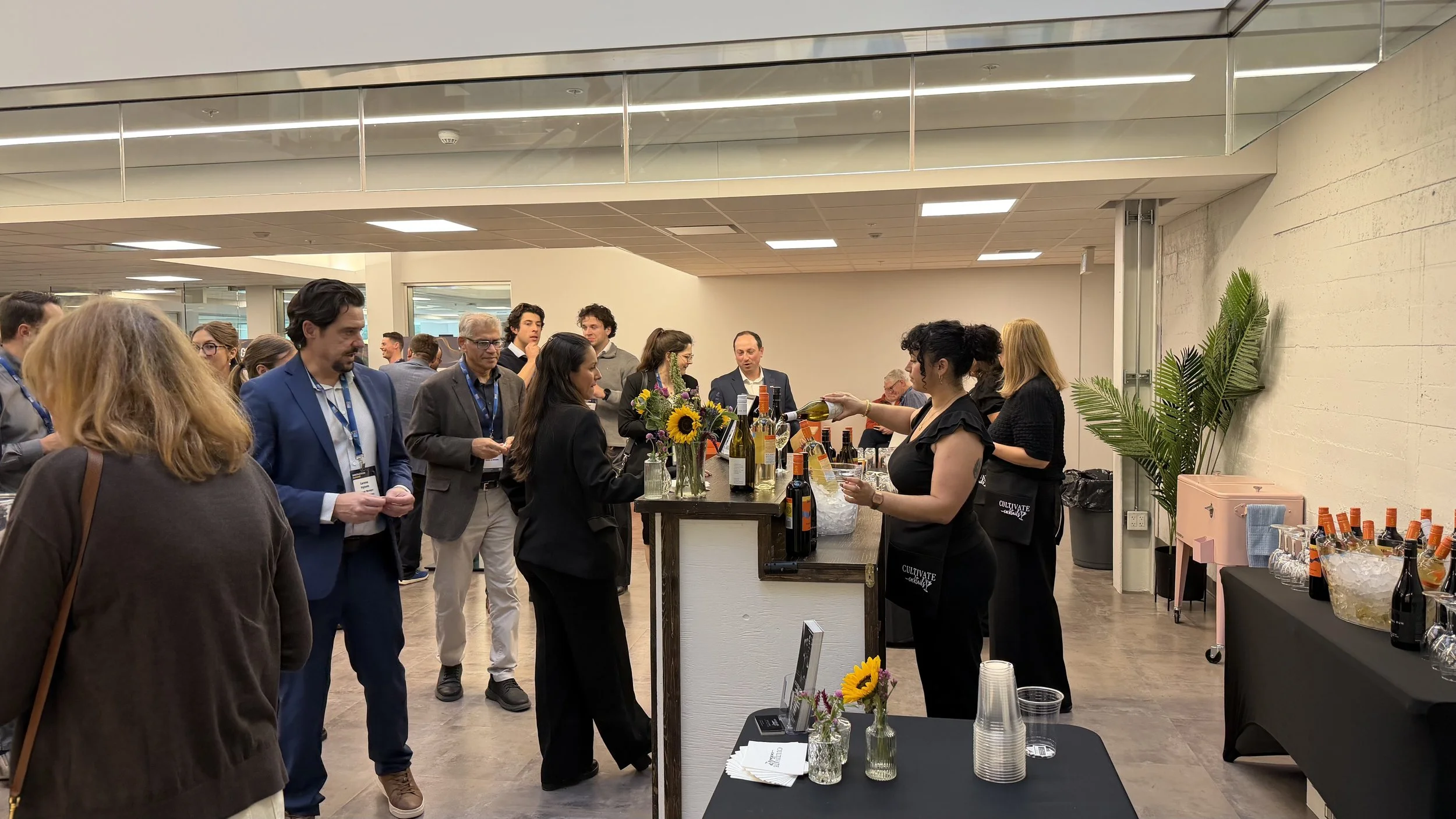 A group of people gathered at a wine tasting event, with a server pouring wine. There are bottles of wine, a sunflower arrangement, and glasses on the table. The setting appears to be indoors with modern decor.