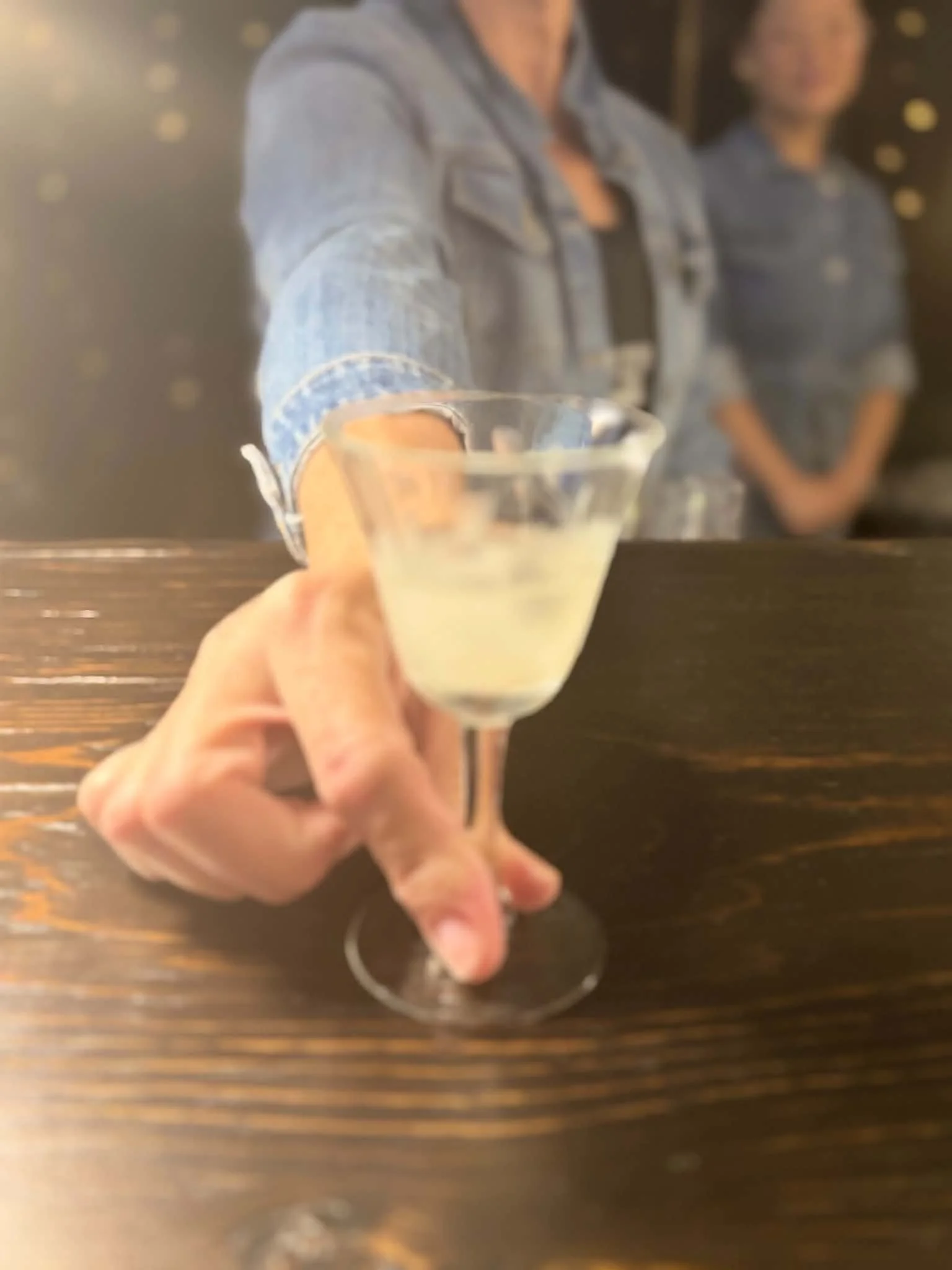 A person holding a glass of a light-colored beverage in front of a mirror with two people reflected behind the glass.