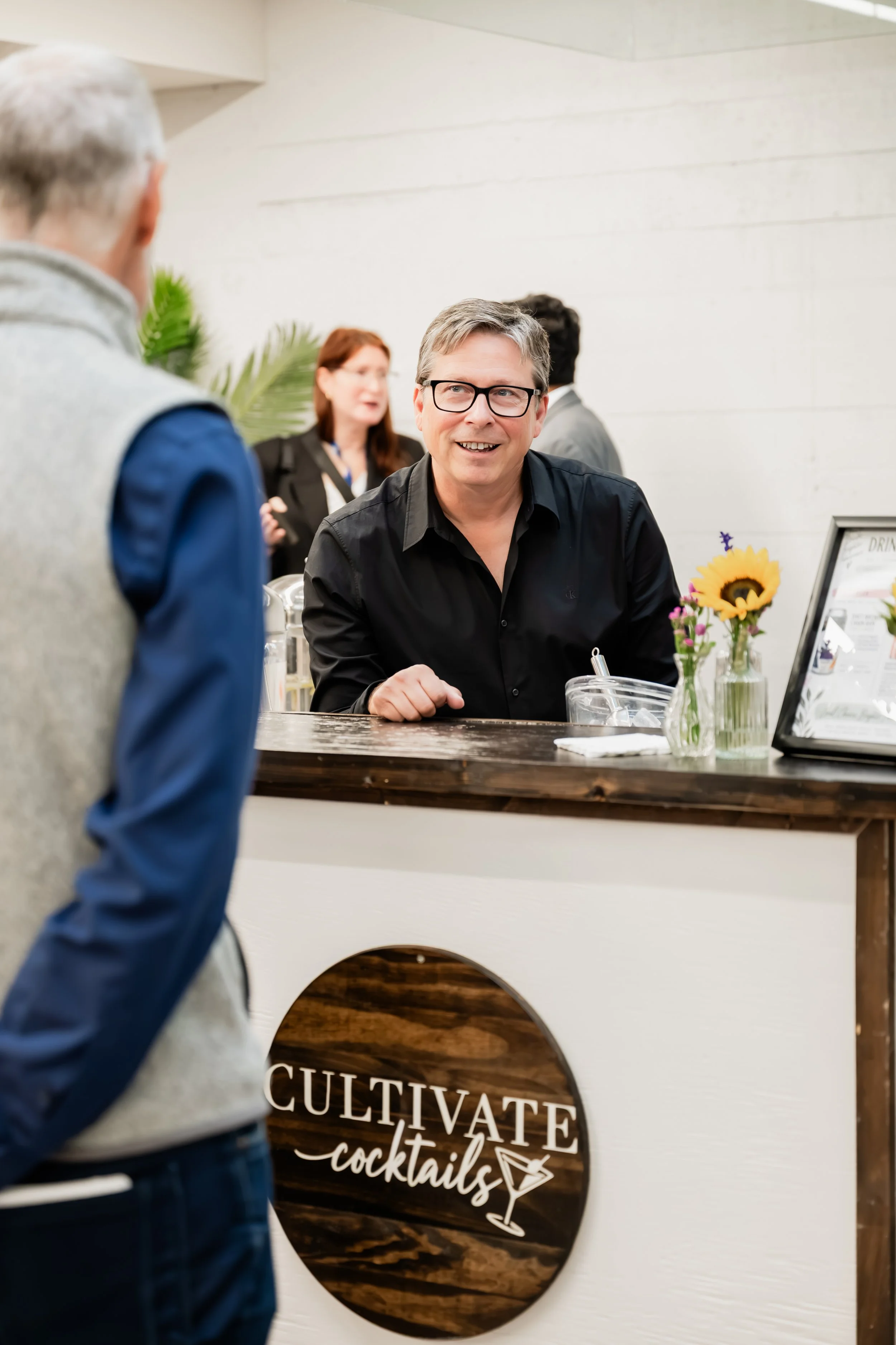 A man with glasses smiling and talking at a counter with a sign labeled 'Cultivate Cocktails' in the background, with people and flowers visible behind.