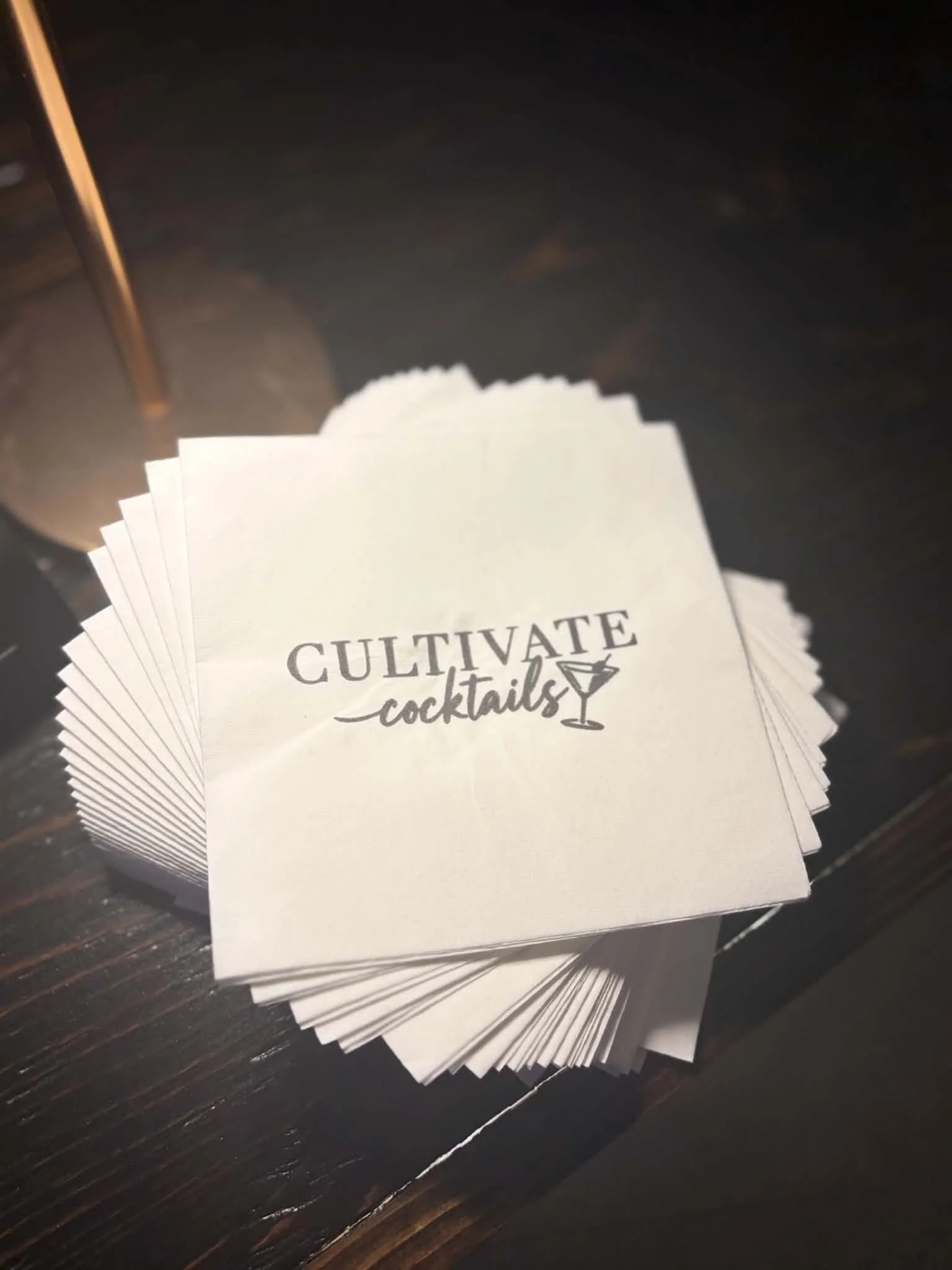 A stack of white napkins with the words 'CULTIVATE cocktails' printed on them, placed on a dark wooden surface.