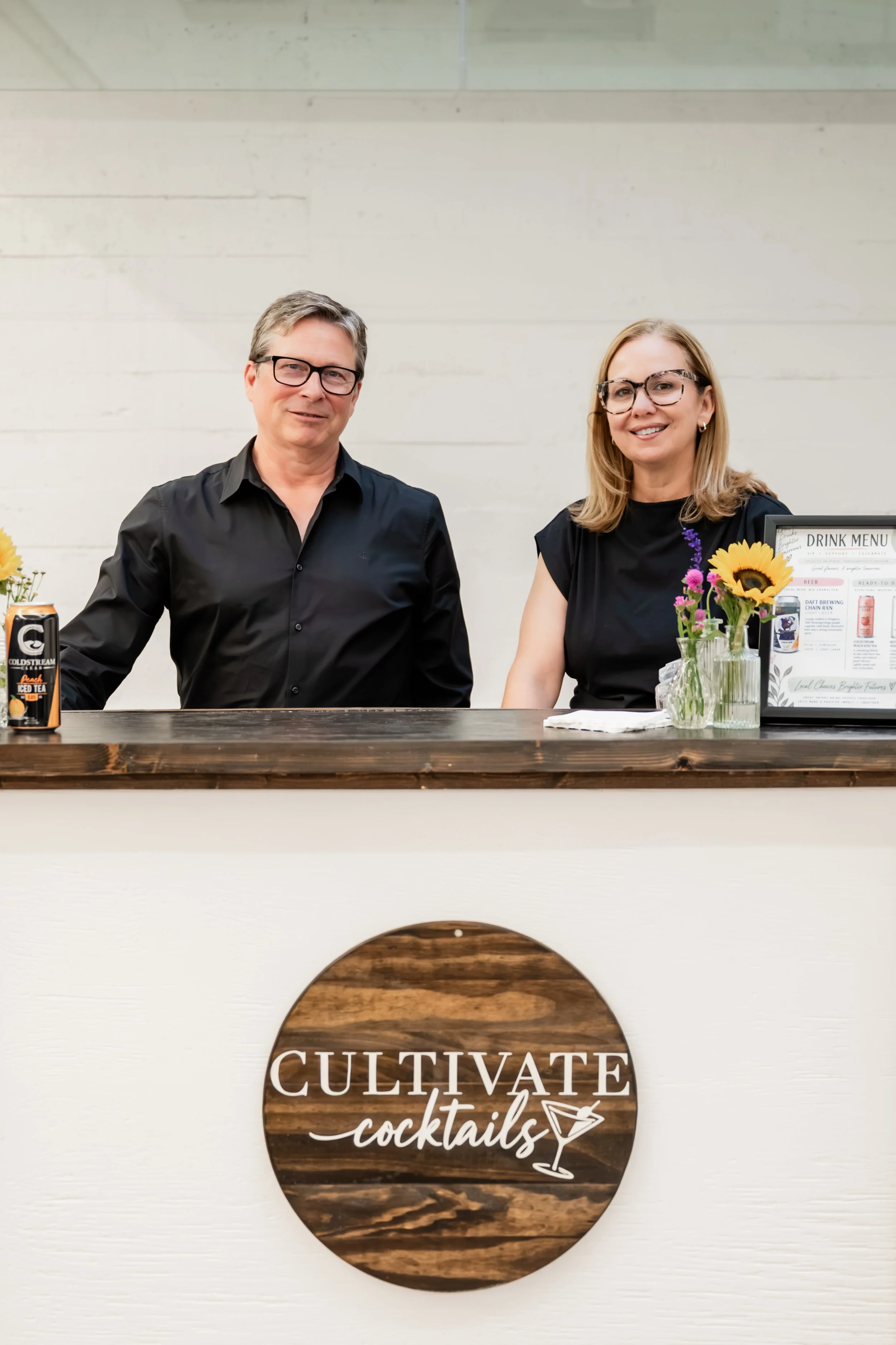 Two people standing behind a bar with a wooden sign that reads 'CULTIVATE cocktails'. There are flowers and a drink menu on the bar. The man on the left wears a black shirt and glasses; the woman on the right wears a black dress and glasses.