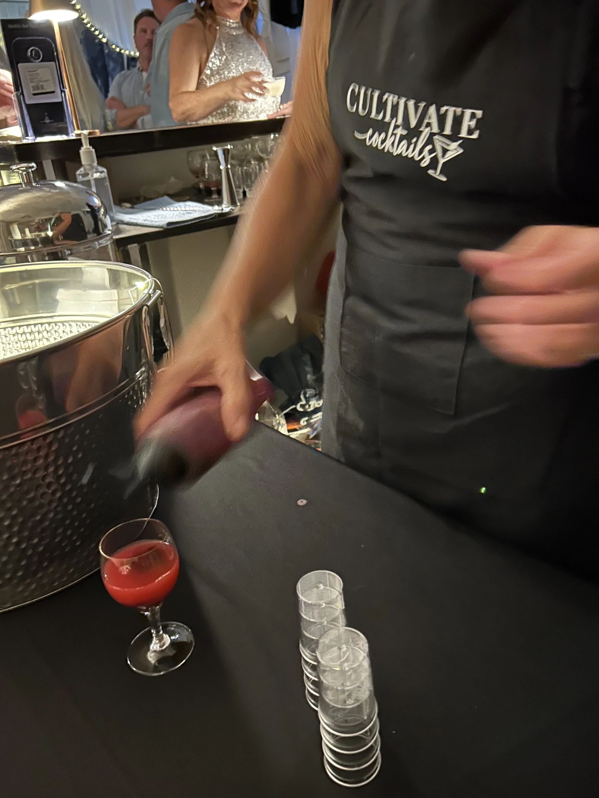 A person in a black T-shirt with white text that reads 'CULTIVATE cocktails' is pouring a red-colored drink into a wine glass. There are clear plastic cups stacked on a black table, and a large glass container with a metal lid on the table. In the ba
