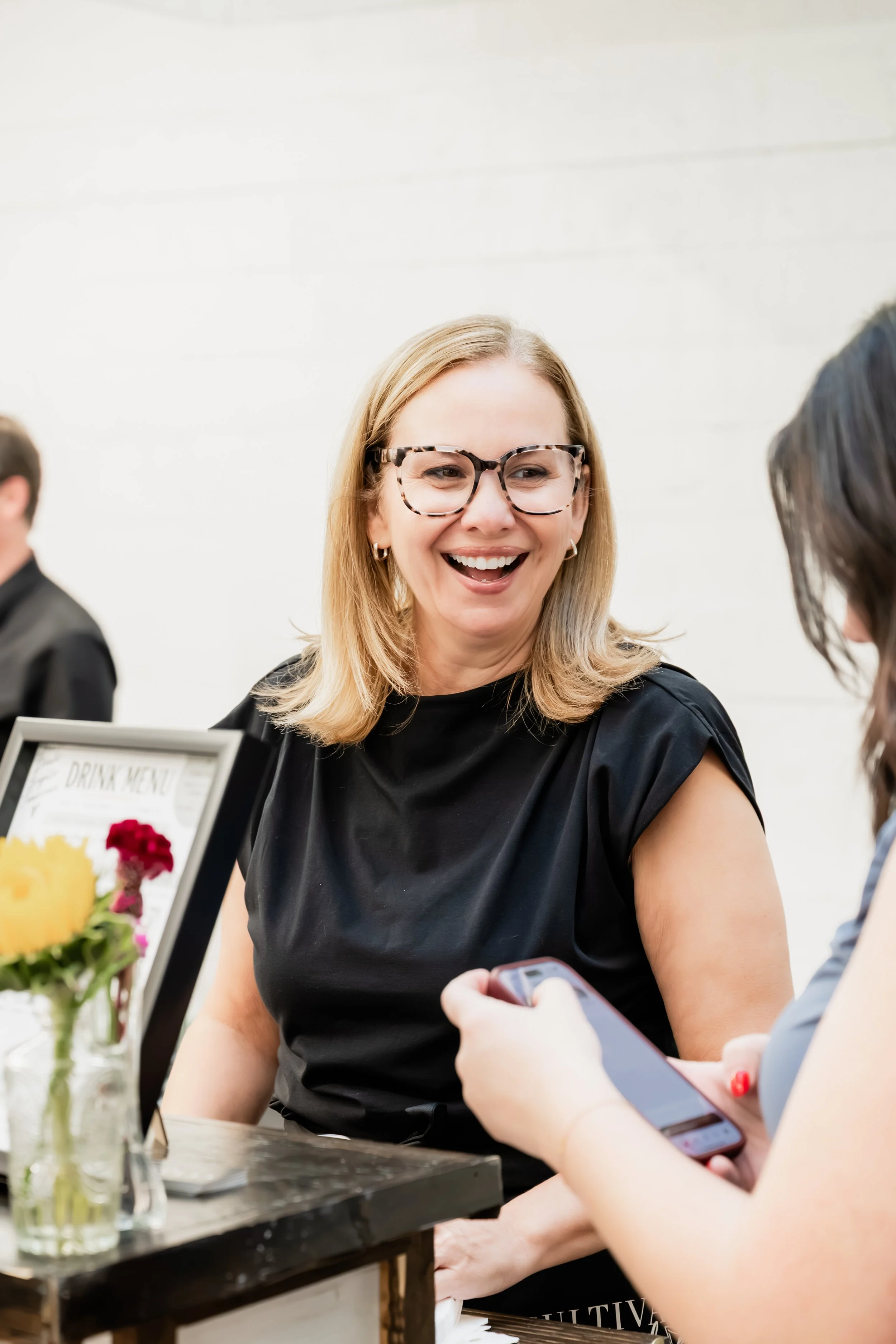 A woman with blonde hair and glasses smiling, sitting at a table, using a smartphone, with friends around her and flowers on the table.