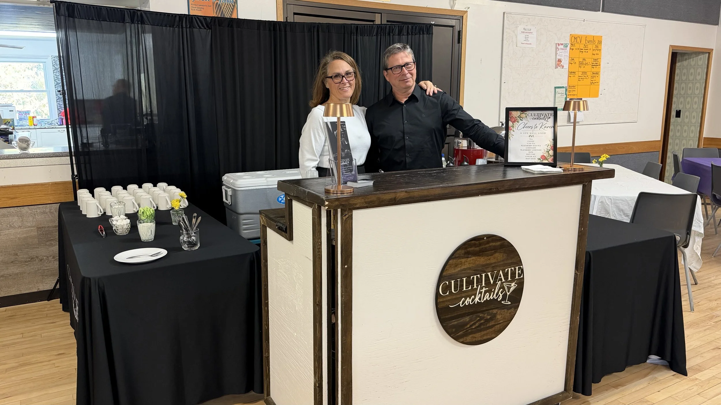 A man and woman standing behind a cocktail bar at an event, with tables set up nearby for guests, and a black curtain backdrop.