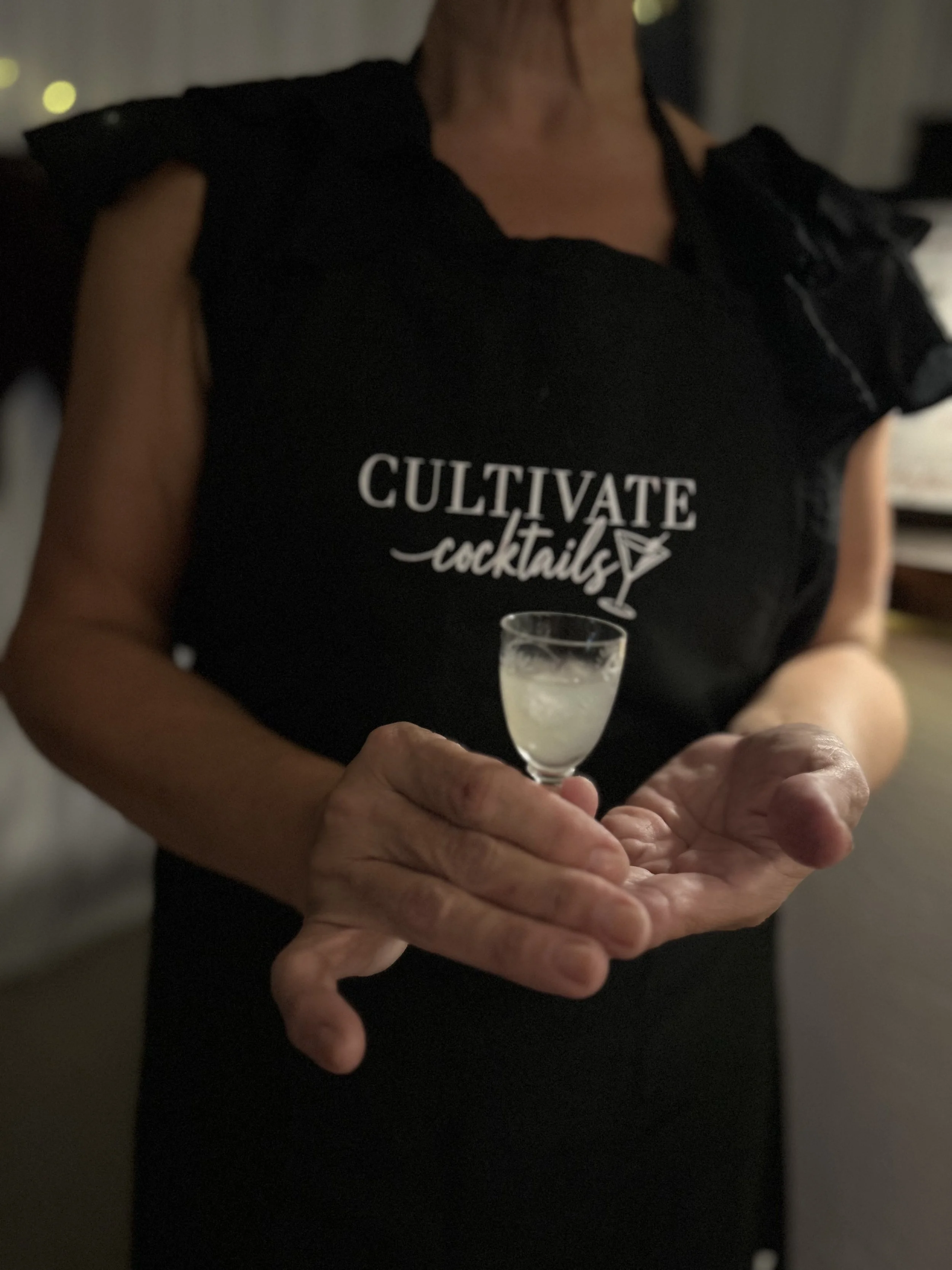 A person holding a small cocktail glass with a light-colored beverage, wearing a black shirt with the words 'Cultivate Cocktails' and a cocktail glass graphic printed on it.