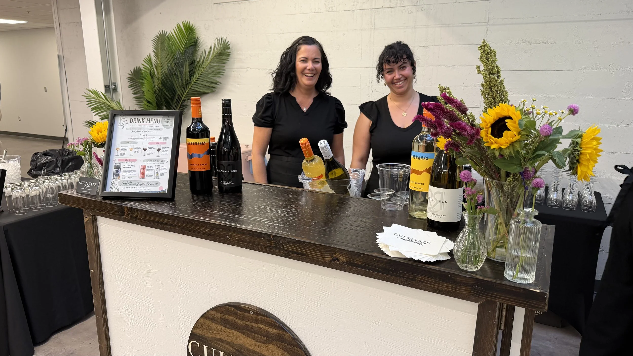 A bar counter with two women smiling, various bottles of wine, a drink menu, and a bouquet of colorful flowers.