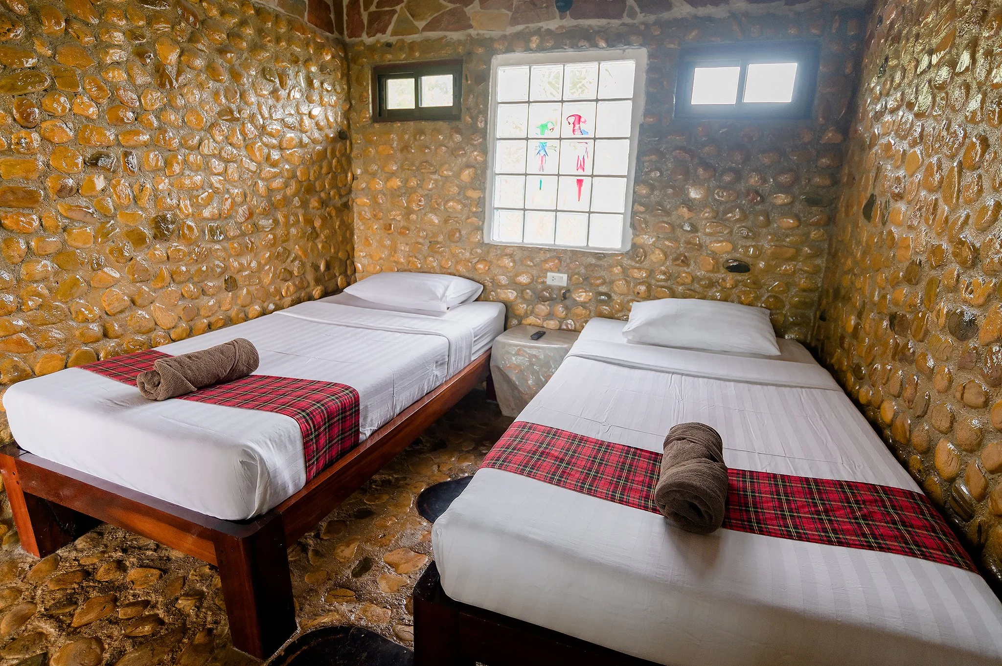 Two twin beds with white linens, rolled towels, and red plaid runners set against stone accent walls and polished tile flooring in a guest room.