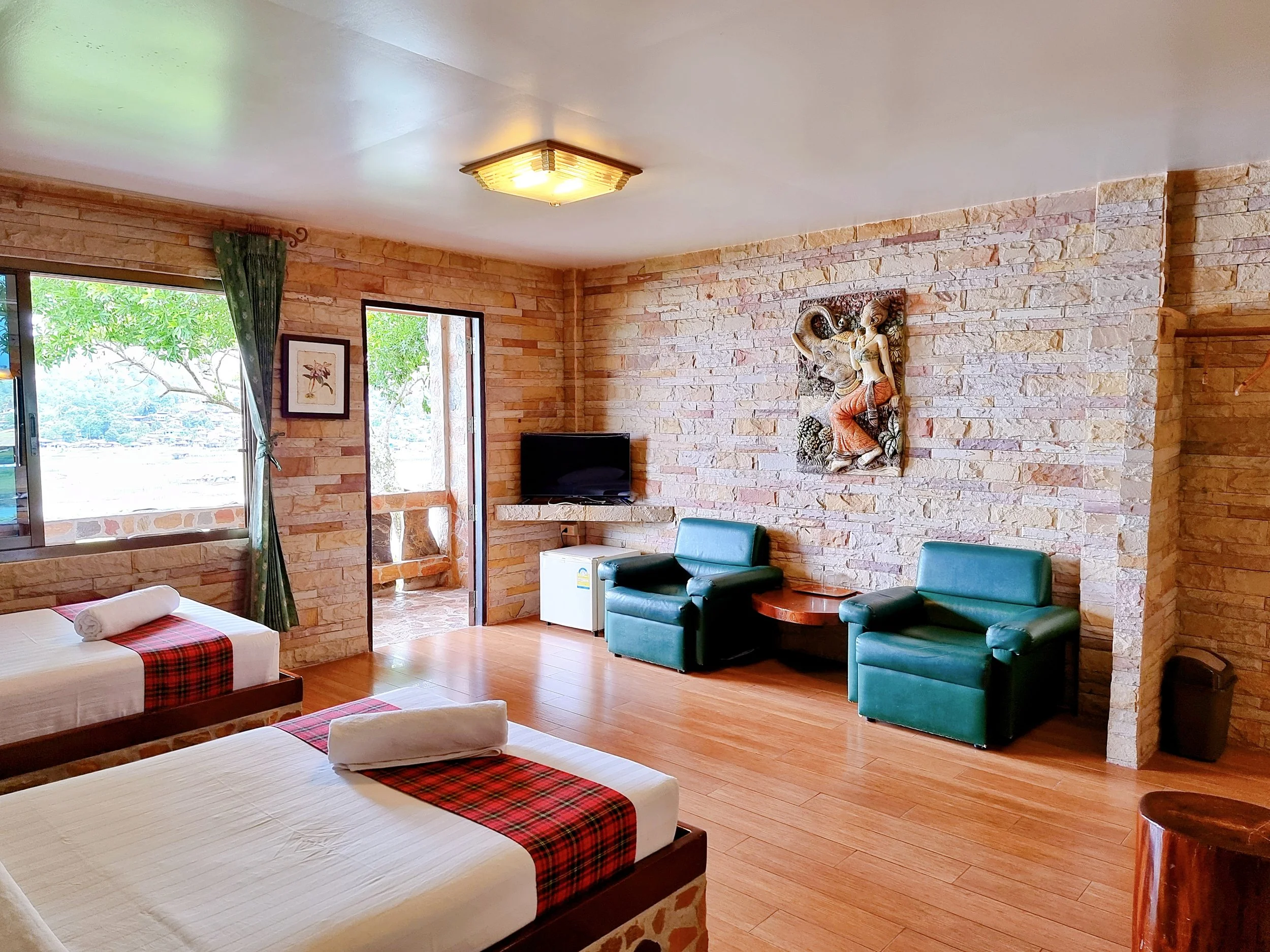 Spacious guest room with two beds with red and green plaid runners, two dark teal armchairs, stone accent walls, a small flat-screen TV, and decorative traditional Thai wall art.