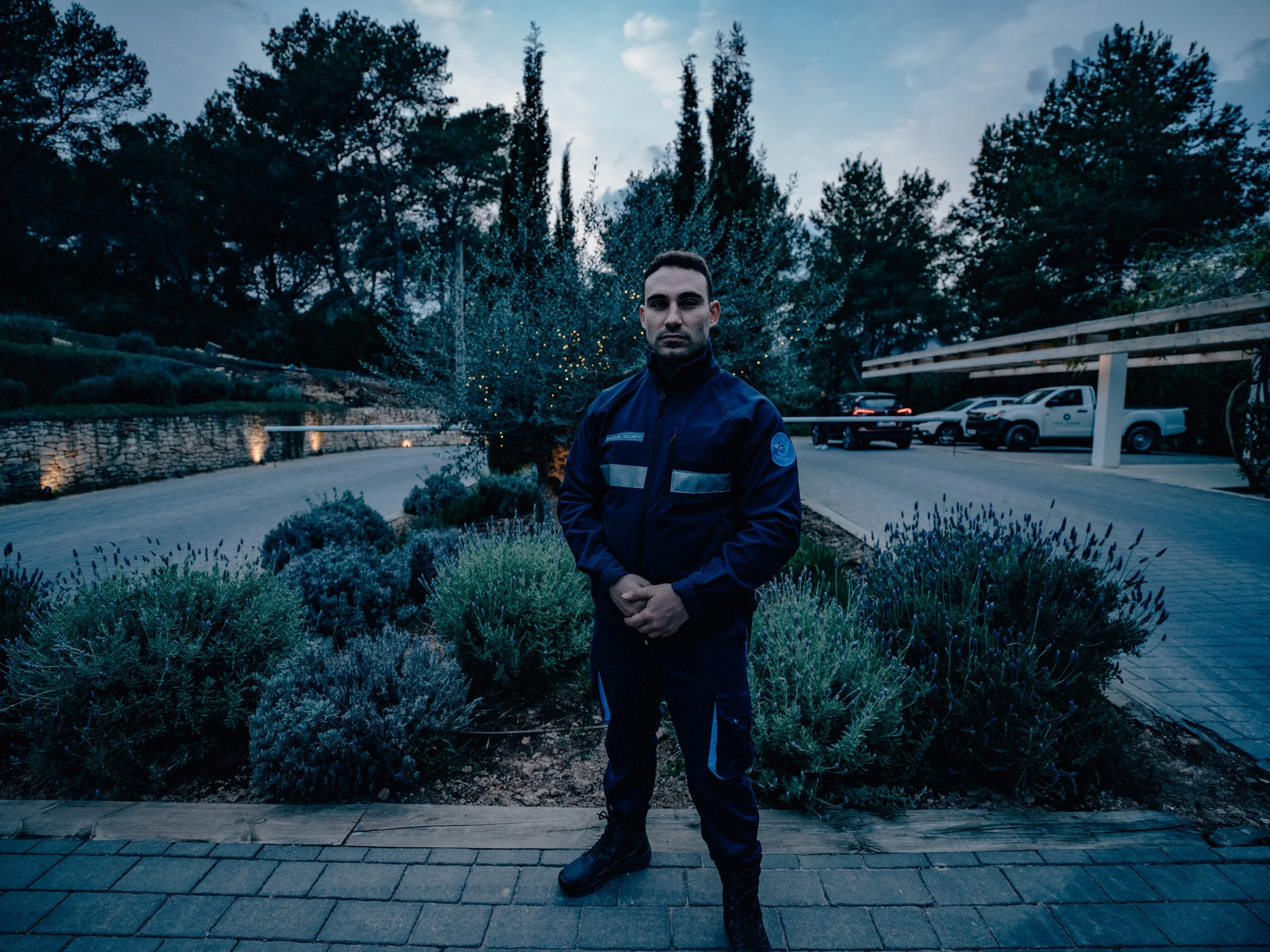 Un oficial de seguridad vestido con uniforme oscuro frente a un arbusto decorativo, con árboles y vehículos en el fondo, en un entorno exterior durante el atardecer o noche.