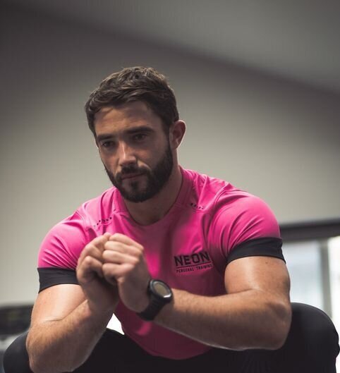 Bearded muscular man in a bright pink athletic shirt sitting indoors with hands clasped.