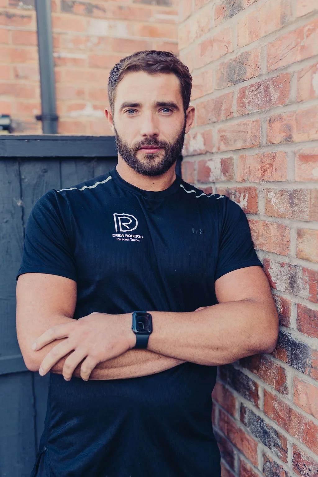 Bearded personal trainer in a black Drew Roberts shirt standing with folded arms against a brick wall