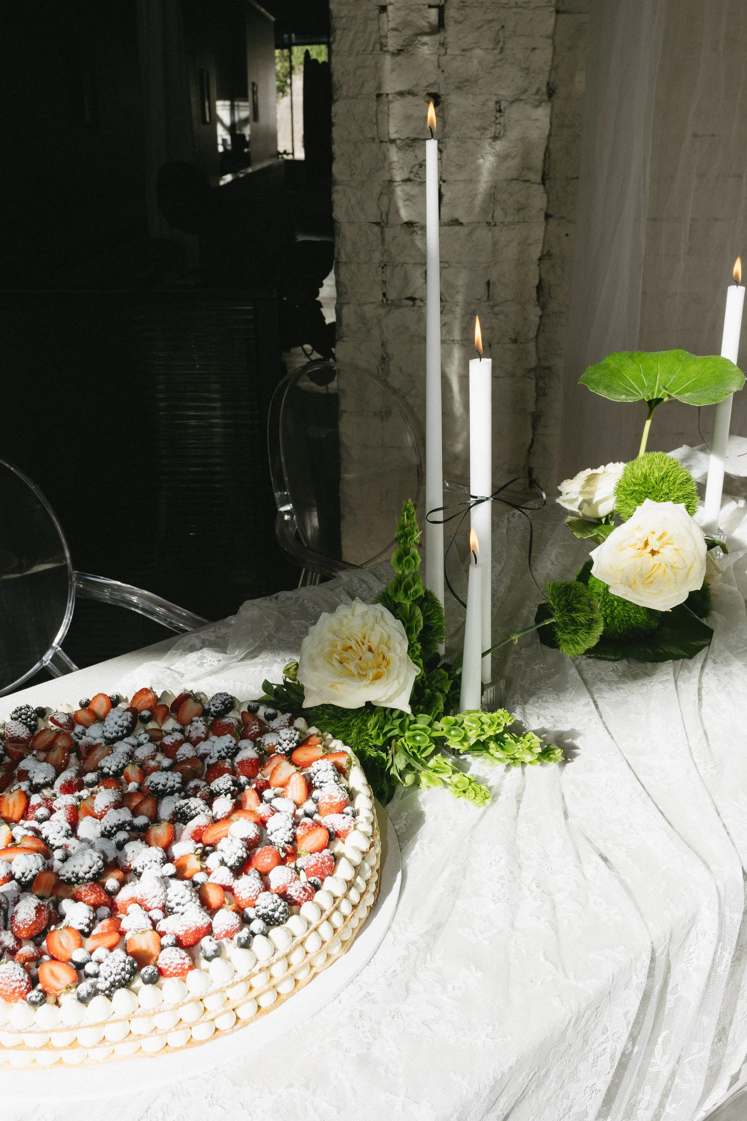 A cake topped with strawberries, blackberries, blueberries, and powdered sugar on a decorated table with white candles and large white flowers.