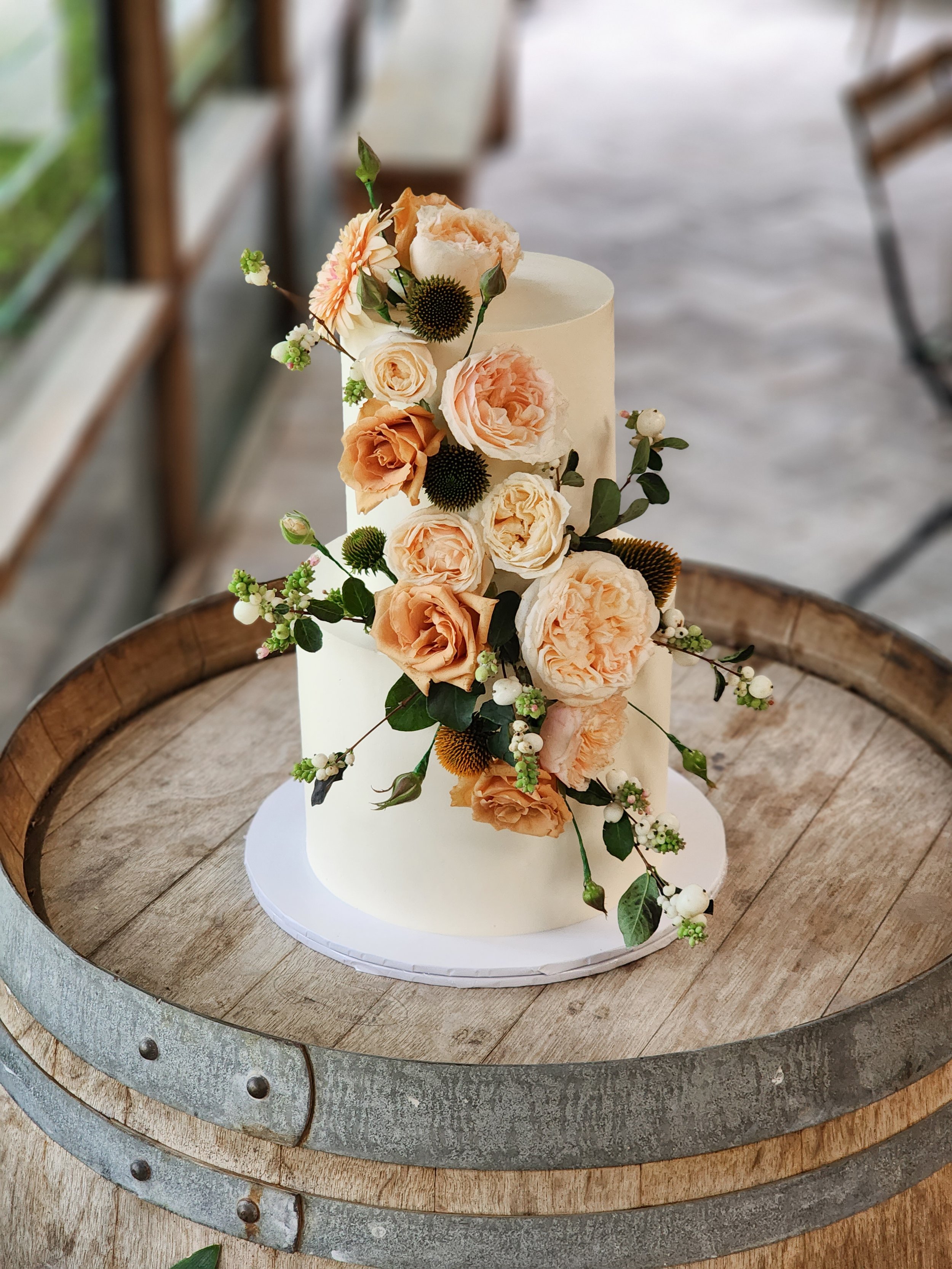 A tall, white wedding cake decorated with cascading peach and cream-colored roses, green leaves, and small white berries, displayed on a round wooden barrel table.