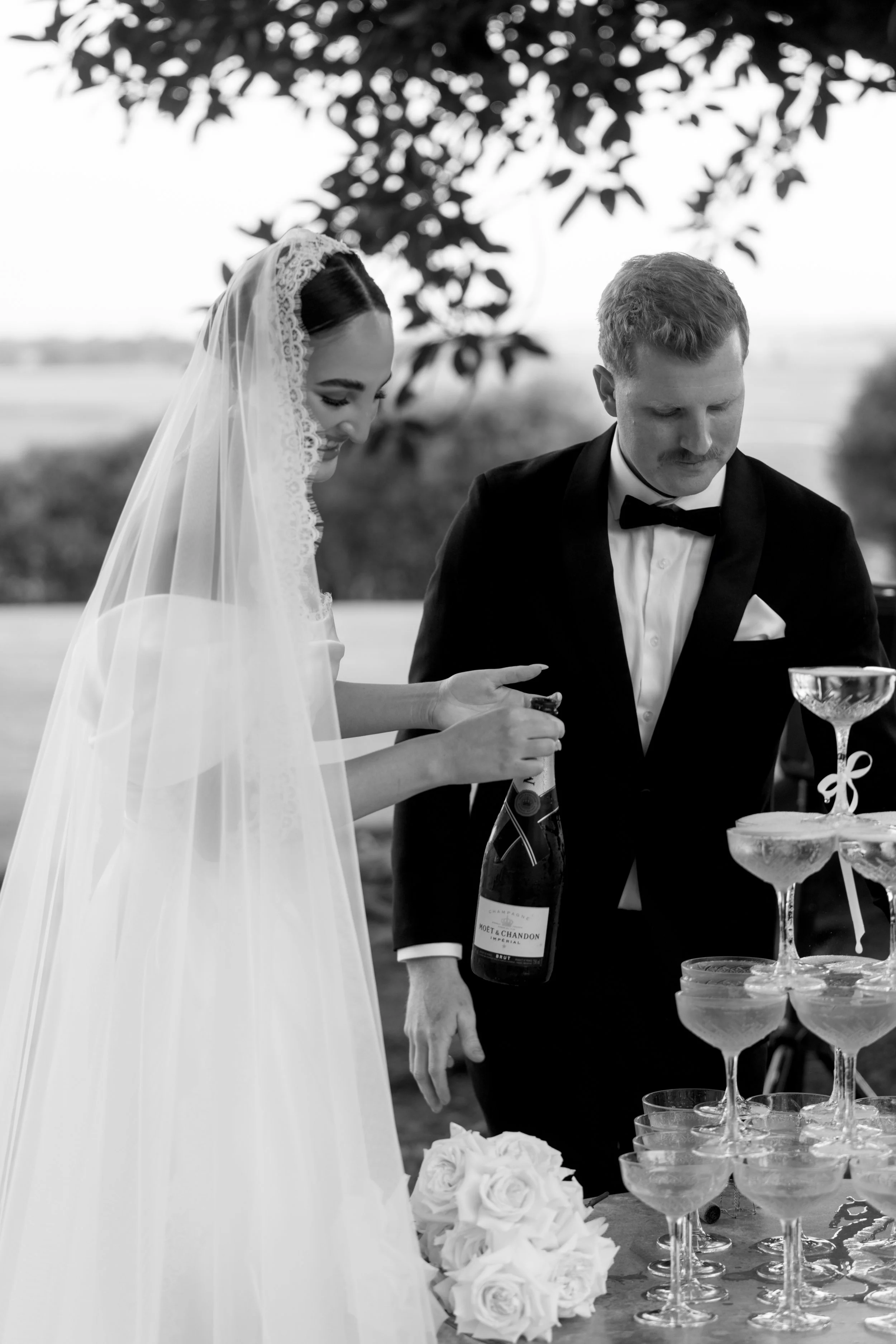 Bride and groom pouring champagne into a tower of glasses, surrounded by roses, at an outdoor wedding.