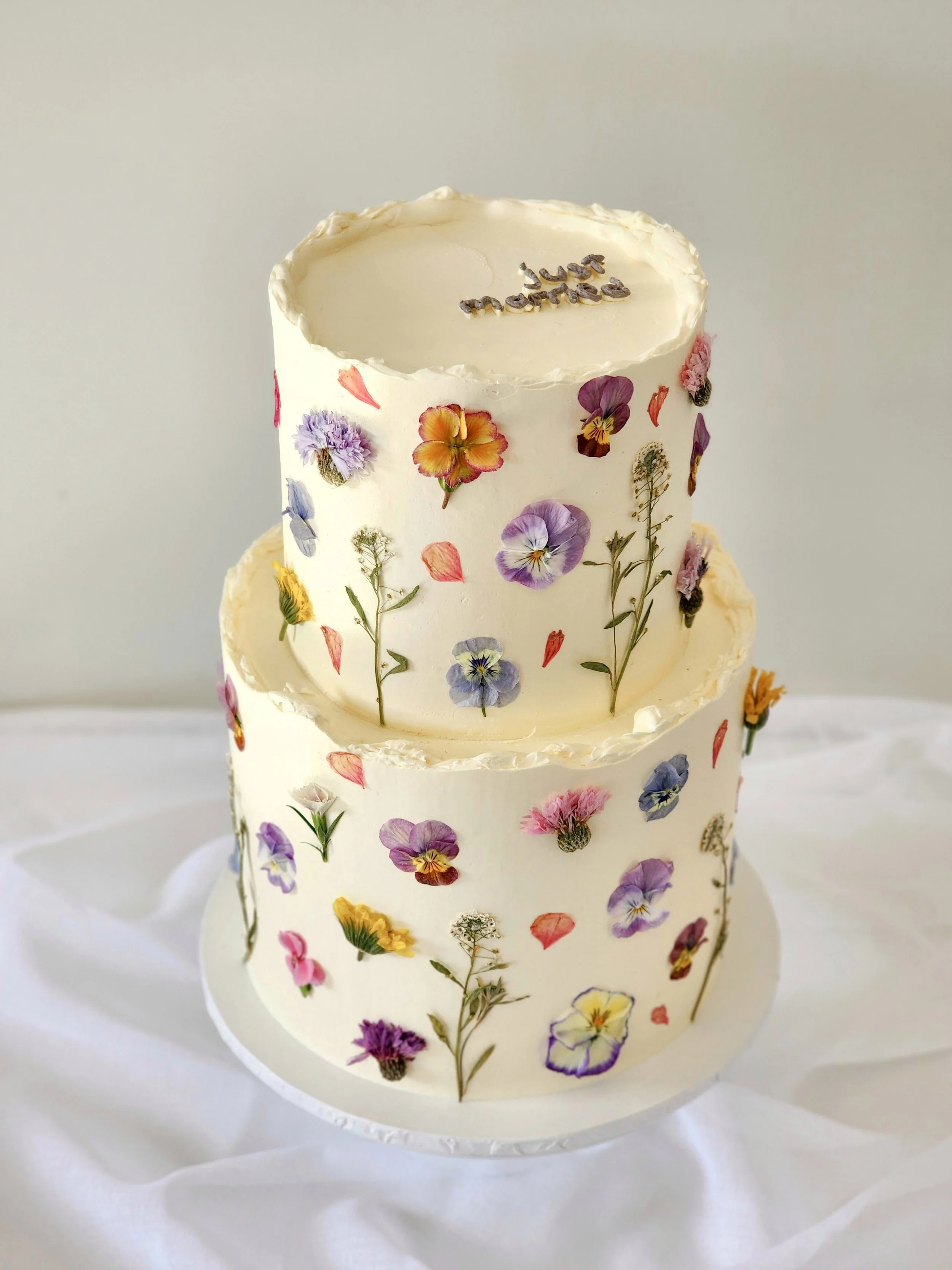 Two-tier white wedding cake decorated with colorful pressed flowers and "just married" on top, on a white tablecloth.