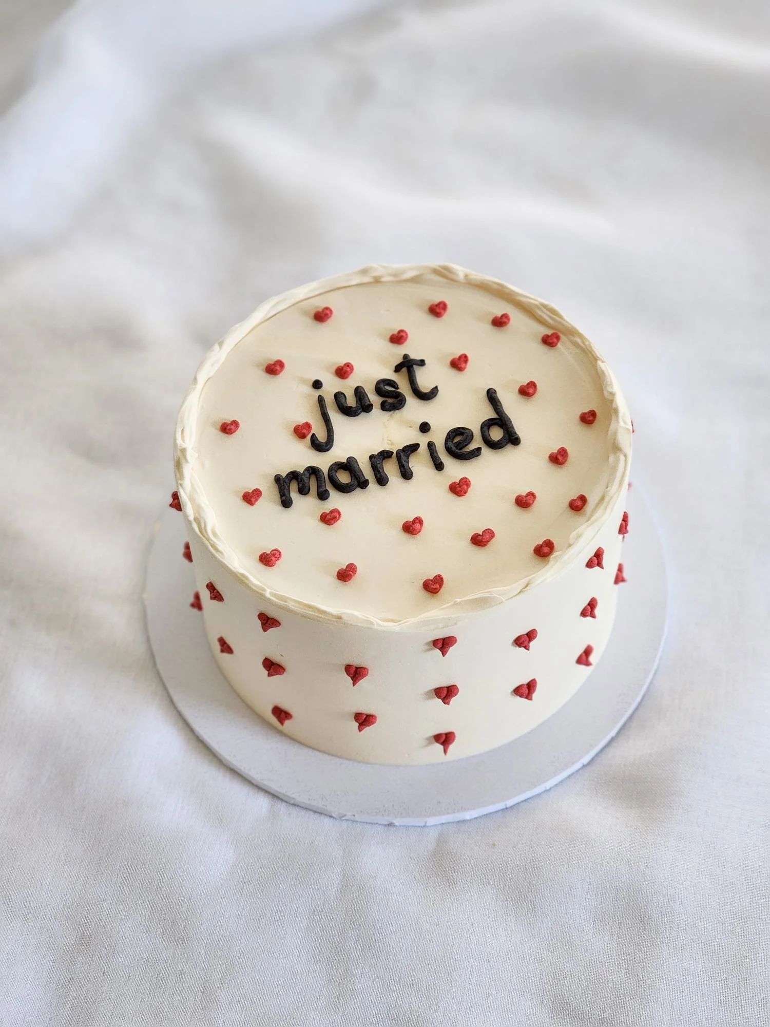 Round wedding cake with white frosting, decorated with small red hearts and the words 'just married' written in black on top.