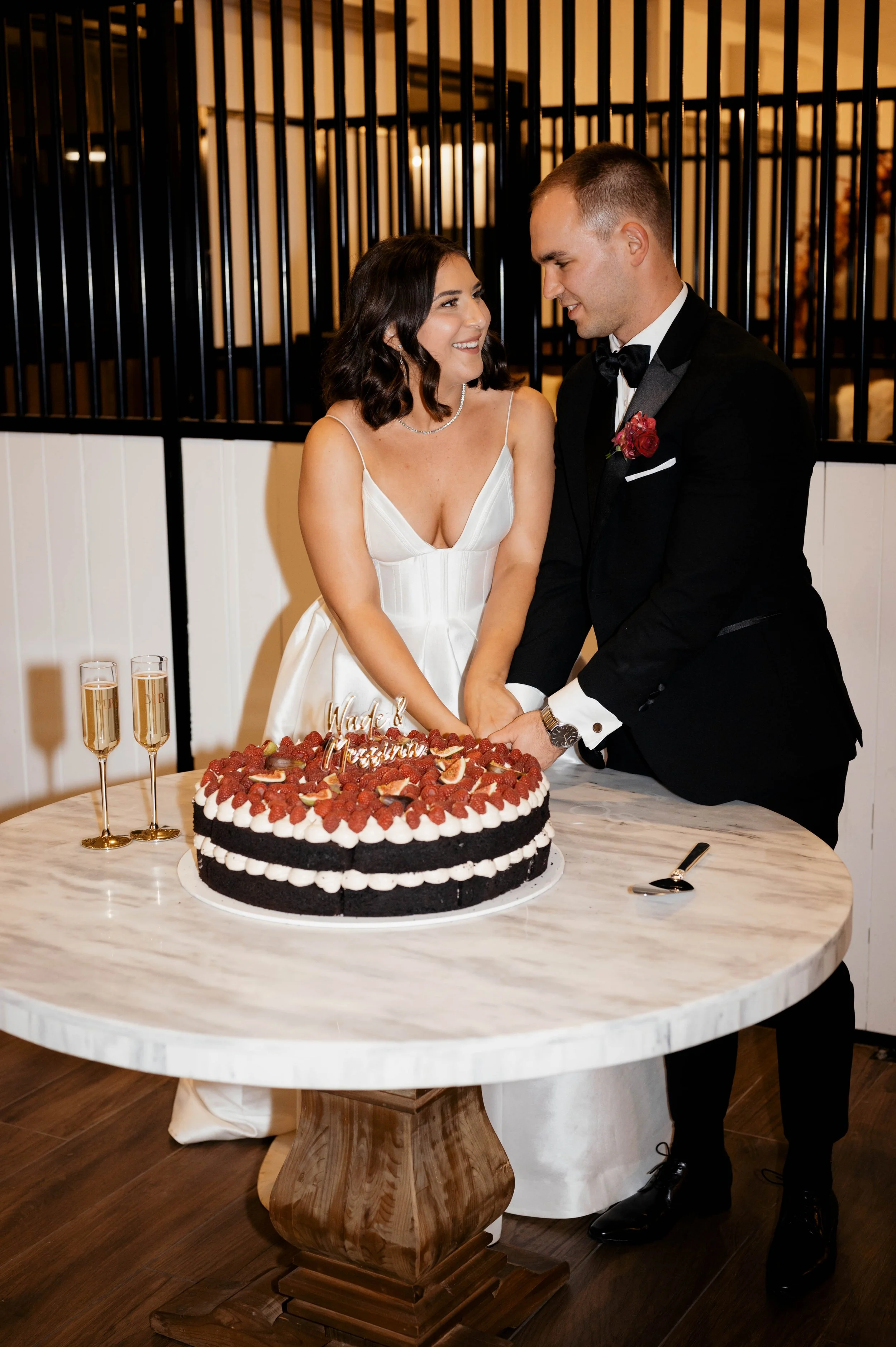A couple cutting a wedding cake. The bride is in a white gown and the groom is in a black tuxedo. The cake is decorated with raspberries and figs, with two champagne glasses nearby on a marble table.
