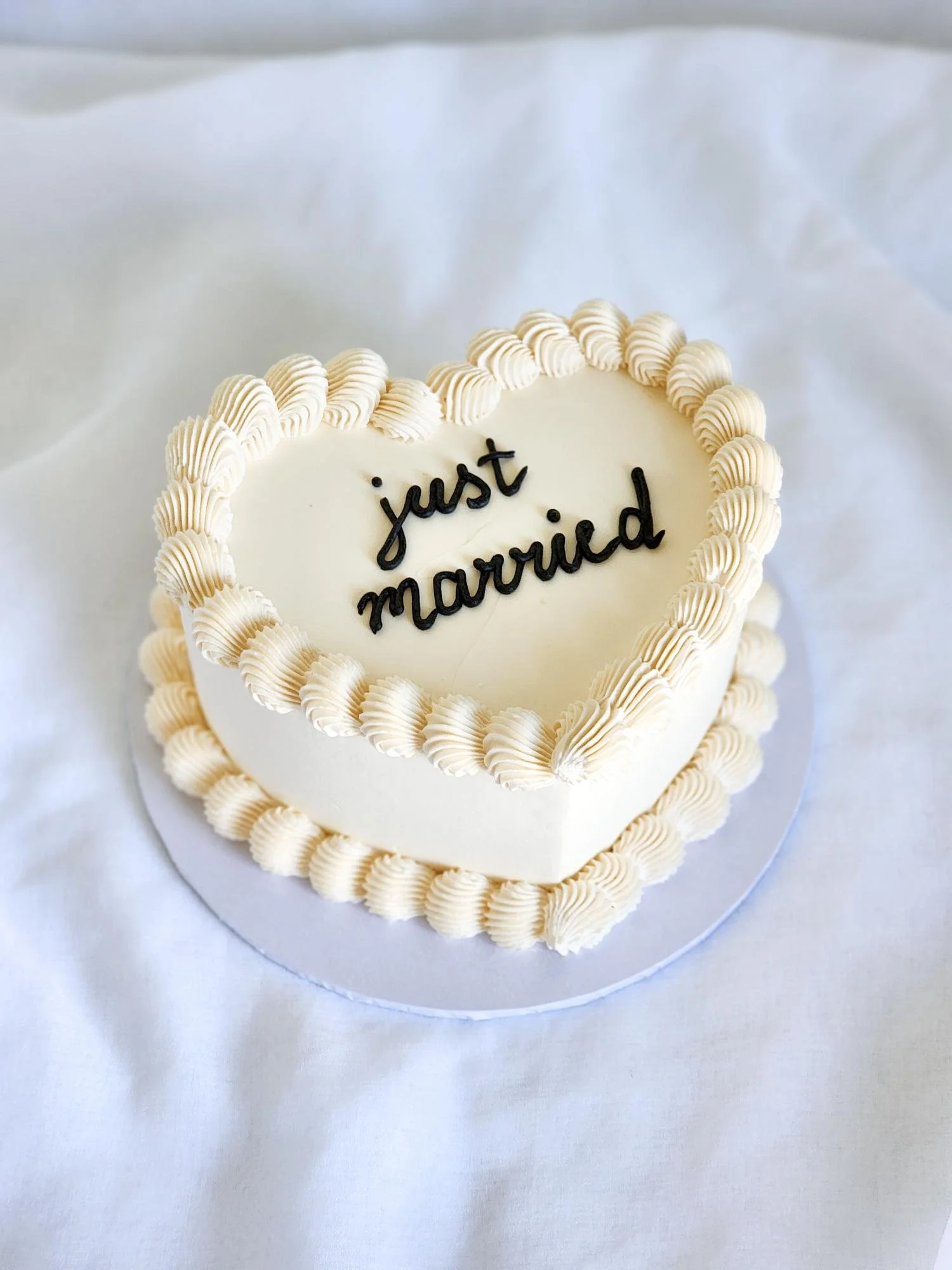 A heart-shaped wedding cake with white frosting and piped border, decorated with the words 'just married' in black icing on top.
